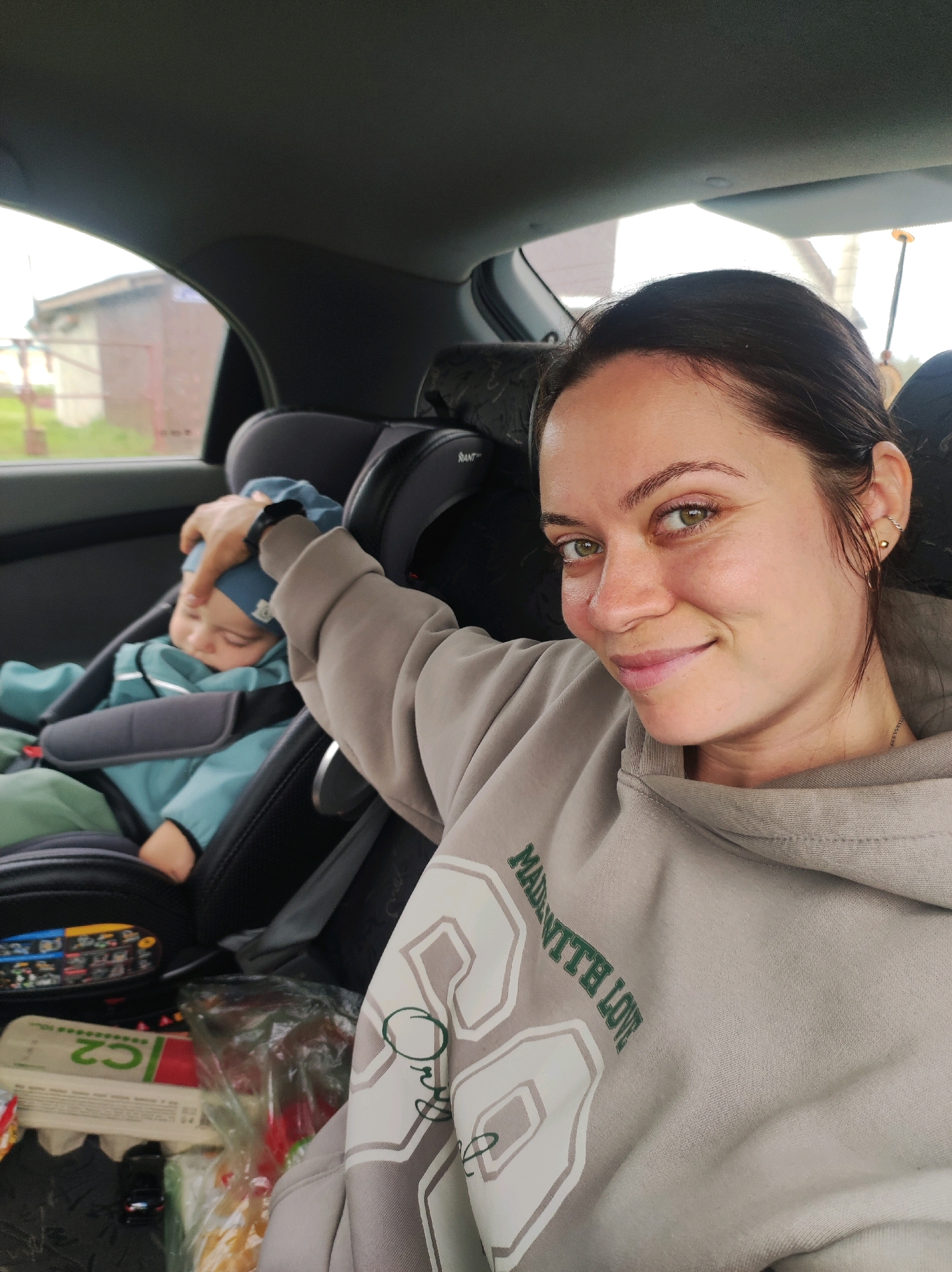 A woman with dark hair and green eyes smiles at the camera from the front passenger seat of a car. She is wearing a light grey hooded sweatshirt with the text "MADE WITH LOVE" printed on it. Her left arm is resting on a baby in a car seat behind her, gently patting the baby's head. The baby, wearing a blue hat and jacket, is asleep in the car seat. The interior of the car is visible, including the car seat and some items in the back, such as a toy car and a plastic bag. The background outside the car window shows a building and some greenery. The image appears to be taken in the city of Fokino, Russia, based on the provided location context.