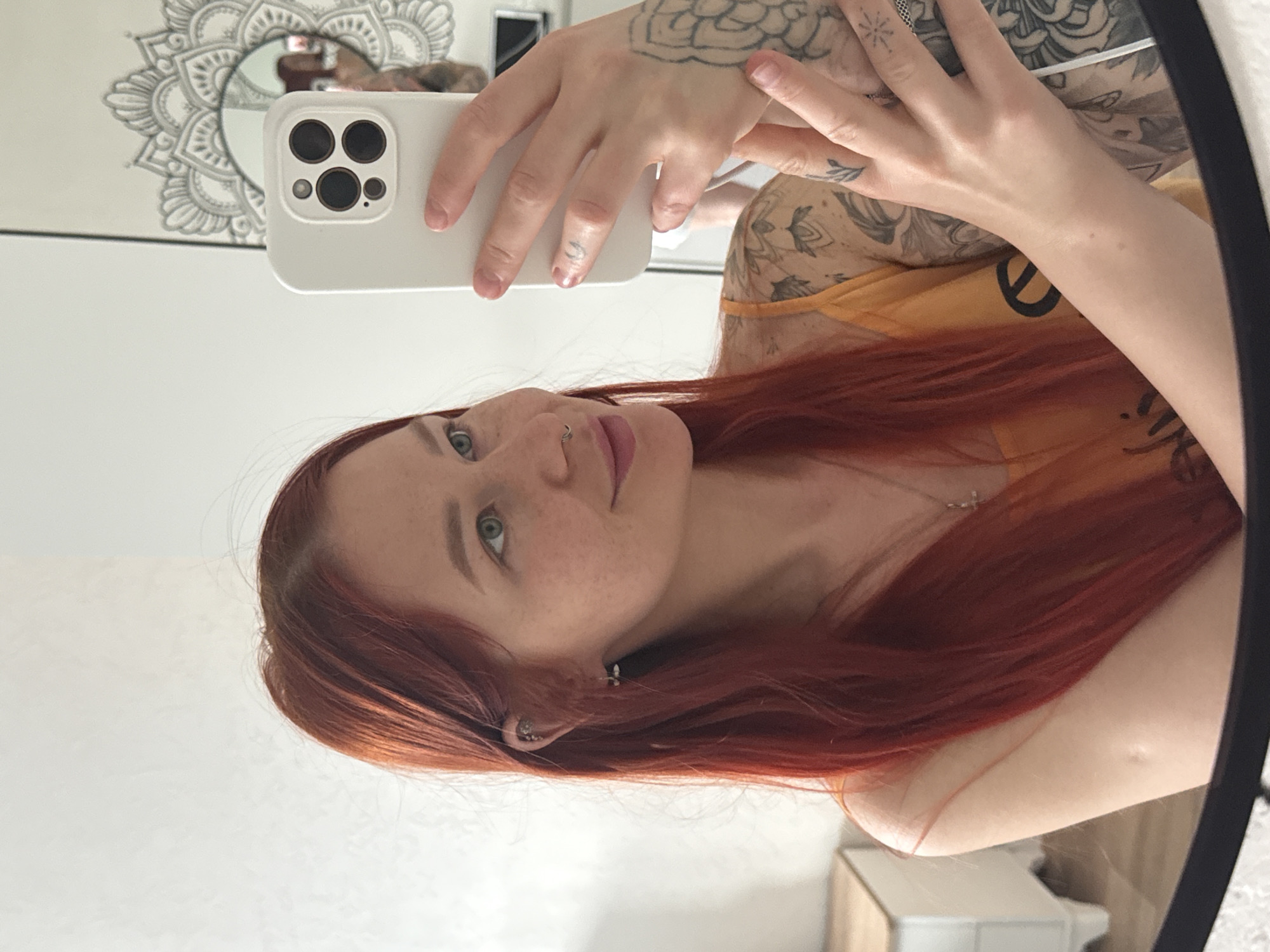The image shows a woman taking a selfie in a mirror. She has red hair and fair skin with freckles. She is wearing a yellow tank top. Her left arm is visible, showing multiple tattoos. She is holding a white smartphone with multiple camera lenses. The background shows a white wall with a decorative mirror and some furniture. I cannot verify the location to be Rostov-on-Don, Russia.