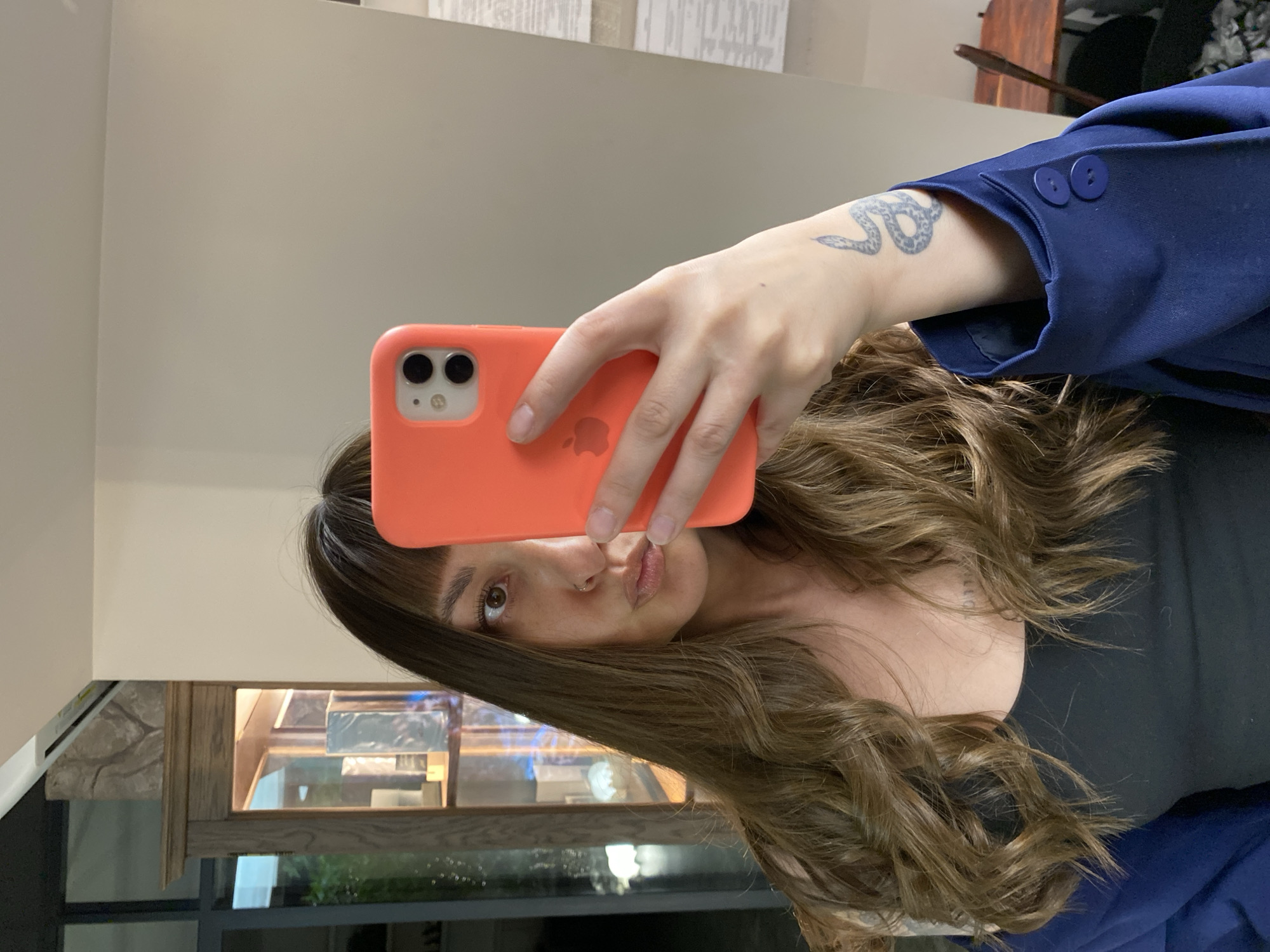 A woman is taking a selfie with a red iPhone. She is wearing a dark blue blazer and a black top. The background includes a wall, a wooden cabinet with glass doors, and a window. There is a snake tattoo on her left arm. The location is possibly indoors, but there is no confirmed information on whether the image was taken in Votkinsk, Russia.