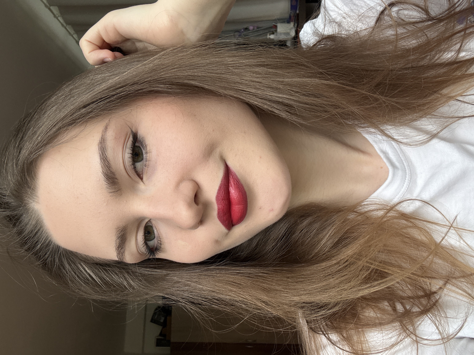 A fair-skinned woman with hazel eyes and long, light brown hair is facing the camera with her head tilted slightly to the right. She is wearing red lipstick and a white t-shirt. Her left hand is touching her hair near her forehead. The background is blurry, but appears to be an interior space, possibly a room in Saint Petersburg, Russia. The lighting appears to be natural daylight.