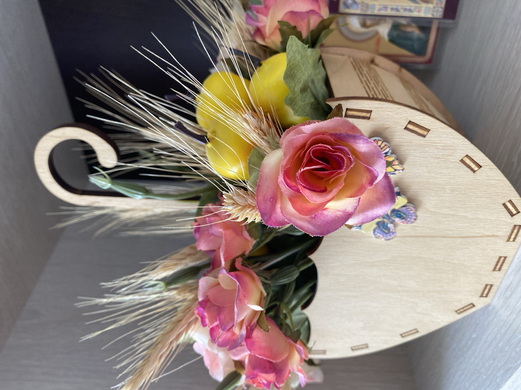 A wooden basket shaped like an umbrella handle holds a bouquet of pink and yellow roses, along with dried wheat stalks and yellow decorative fruit. The basket is adorned with small, colorful butterfly cutouts. In the background, a shelf holds religious icons. The scene appears to be an indoor setting, possibly a home in Lida, Belarus.