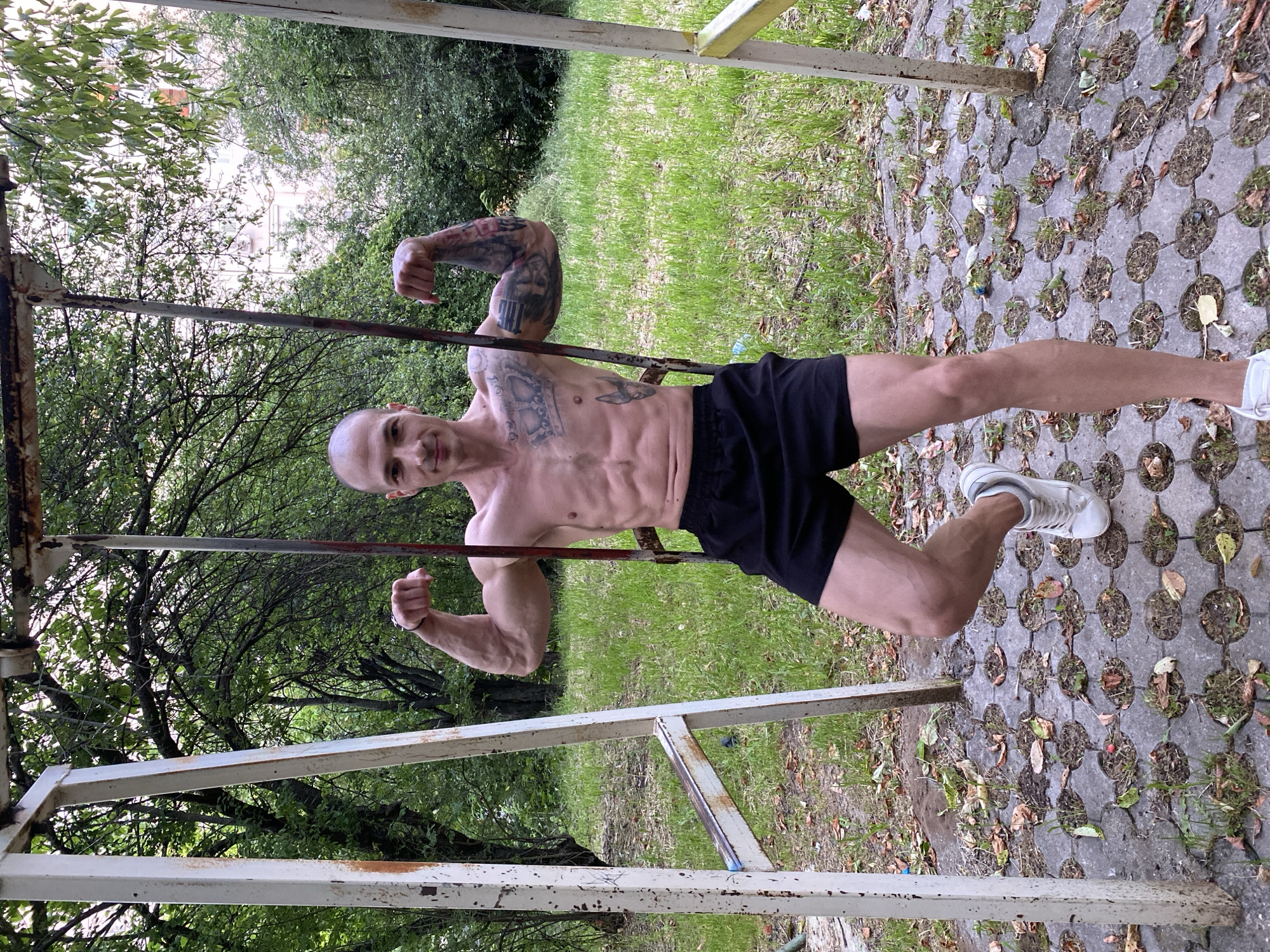 A shirtless man in black shorts and white sneakers poses on an outdoor exercise structure in Pleven, Bulgaria. The man has a shaved head and visible tattoos on his chest and left arm. He holds his arms bent, flexing his biceps. The exercise structure consists of metal bars and posts, some with rusted surfaces. The ground is covered in patterned concrete pavers with grass and leaves filling the gaps. In the background, green trees and a grassy area are visible.