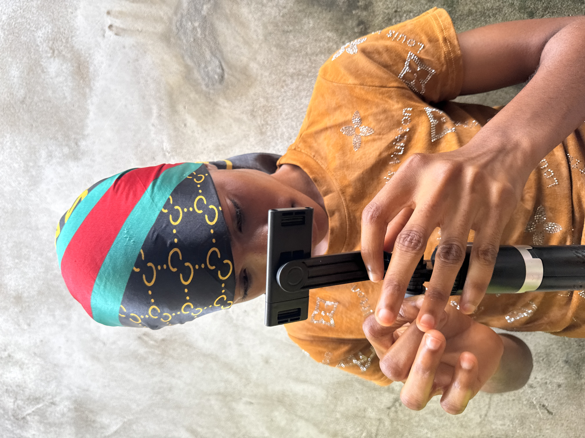 An individual with a dark complexion is holding a black stabilizer in their hands. The person is wearing a yellow shirt adorned with a repeating pattern of stylized four-petal flower designs formed by small crystals. The individual has a headscarf of black fabric with a repeating gold "C" pattern and stripes of red and green. The background appears to be a textured grey surface. There is no verifiable evidence to support the image was taken in Badagry, Nigeria.