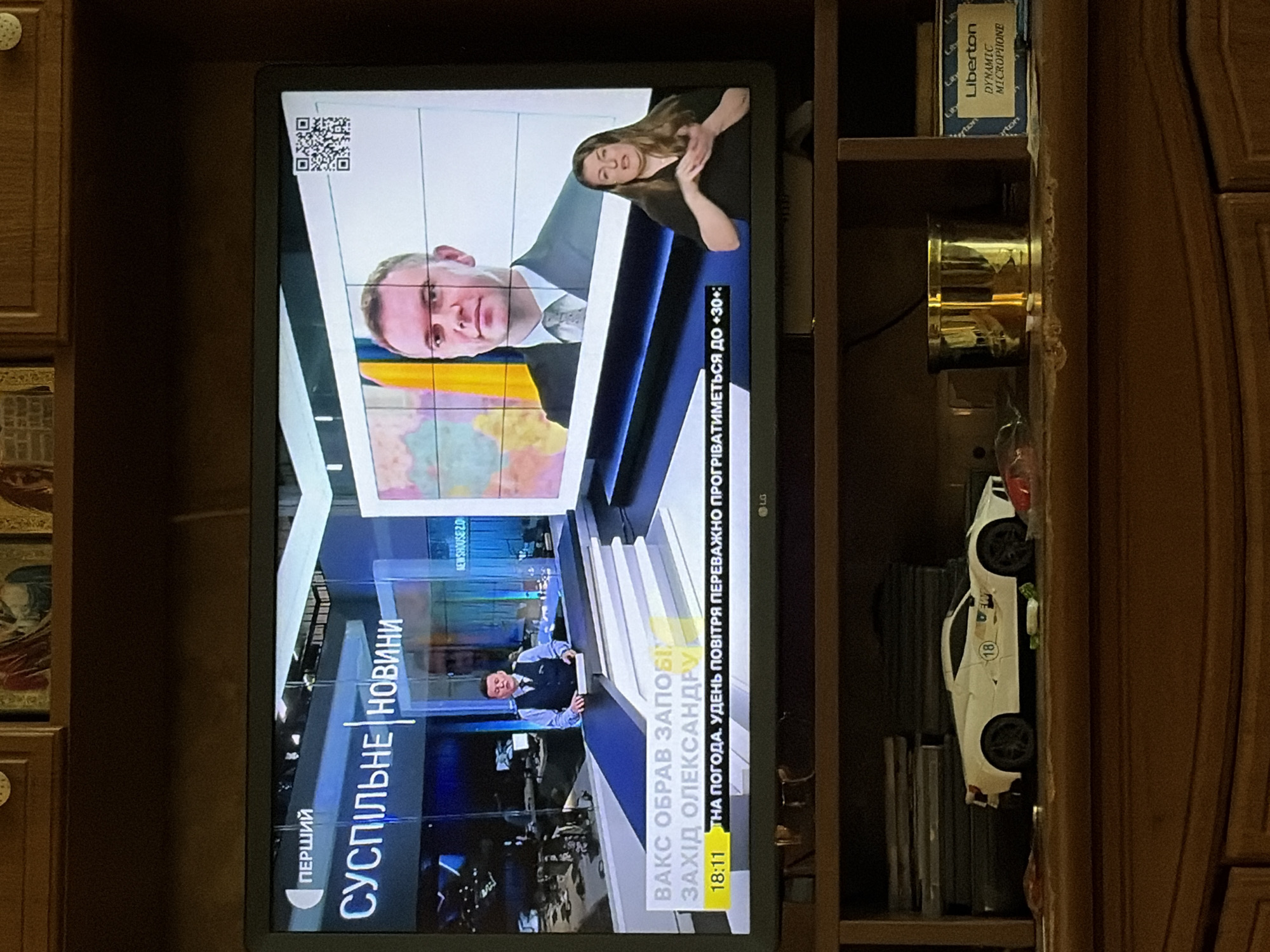 The image shows a television screen displaying a news broadcast. The broadcast features two presenters, one male and one female, who appears to be a sign language interpreter. The male presenter is in the upper half of the screen, wearing a suit and tie, and speaking at a desk. The female interpreter is in the lower right quadrant of the screen, with her hands raised as if signing. The bottom of the screen displays text information, including the time and weather forecast. The broadcast appears to be in Ukrainian, with the words "ПЕРШИЙ" and "СУСПІЛЬНЕ НОВИНИ" visible. The television is placed on a shelf in a dimly lit room. Below the television, on the shelf, are various objects, including a white toy car, a golden cup, and a box labeled "Liberton DYNAMIC MICROPHONE". The background of the room is not clearly visible, but it appears to be a domestic setting with wooden furniture. The overall scene suggests a casual viewing of a news program at home.
