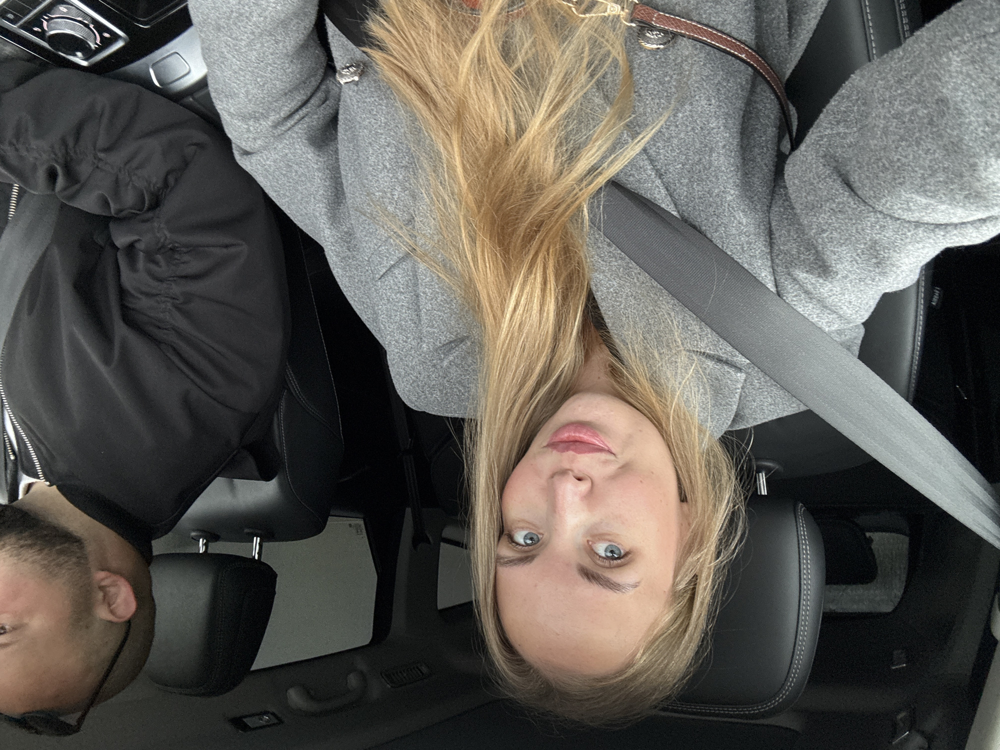 A young woman with blonde hair and blue eyes is in the passenger seat of a car. She is wearing a grey wool coat and a seatbelt. Next to her, in the driver's seat, is a man with a beard and glasses, wearing a black jacket. The interior of the car is black leather. The photo appears to be taken inside a car but there are no visible clues to determine the location within Sochi, Russia.
