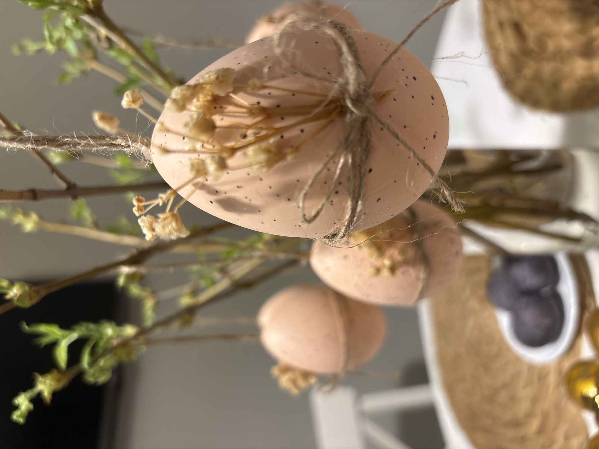 The image shows a close-up of Easter decorations, likely indoors. Three speckled, pale pink Easter eggs are suspended from branches with small green buds and dried flowers. Each egg is tied with a length of twine. The eggs and branches are in focus, while the background is blurred. A woven basket and a white dish containing dark-colored objects are visible in the background. It is not possible to confirm a location in Chaikovsky, Russia based on this image.