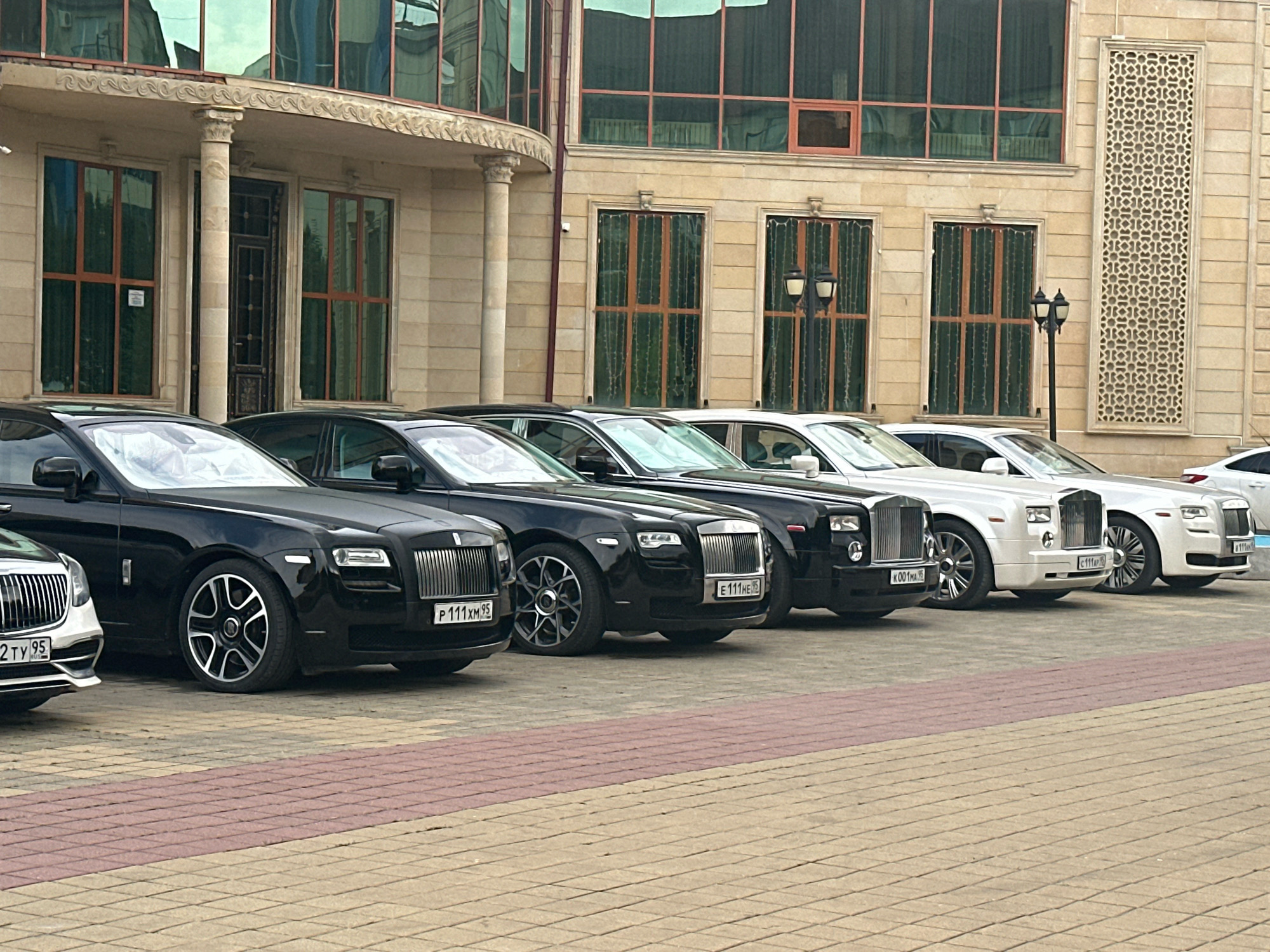 A row of high-end Rolls-Royce vehicles is parked in front of a building in Grozny, Russia. The cars, including black and white models, are lined up on a paved driveway. The building features classical architectural elements with columns, large windows, and ornate decorative panels. The environment appears to be an urban setting, possibly a hotel or private residence. There are no people visible in the image. The scene is static and devoid of any immediate action.