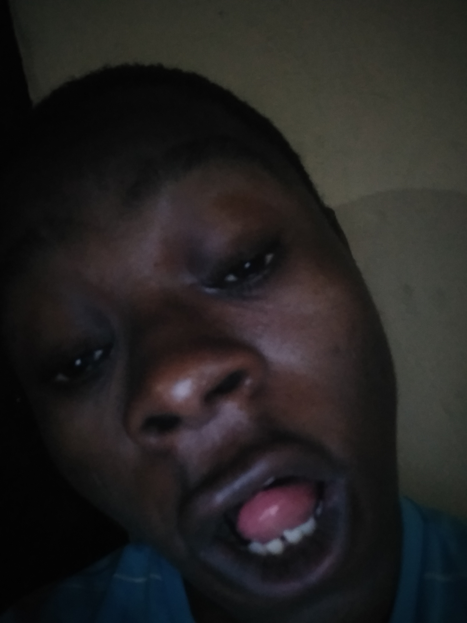 A close-up, low-angle shot of a person with dark skin, presumably male, with their mouth open and tongue visible. The person has a short haircut. The lighting is poor and there are visible wall surfaces behind the person. No location or city can be confirmed.