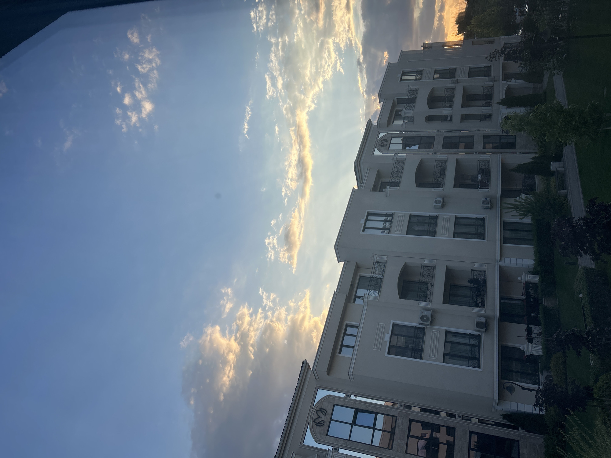 The image captures a modern apartment building in the city of Plovdiv, Bulgaria, bathed in the warm light of a sunset. The building is light-colored, with multiple stories featuring balconies with ornate railings. The sky above is a clear blue, with scattered clouds tinged with orange and yellow from the setting sun, with visible sun rays piercing through them. Green trees and bushes surround the building, with a manicured lawn in the foreground. The architecture of the building suggests it is a residential complex.