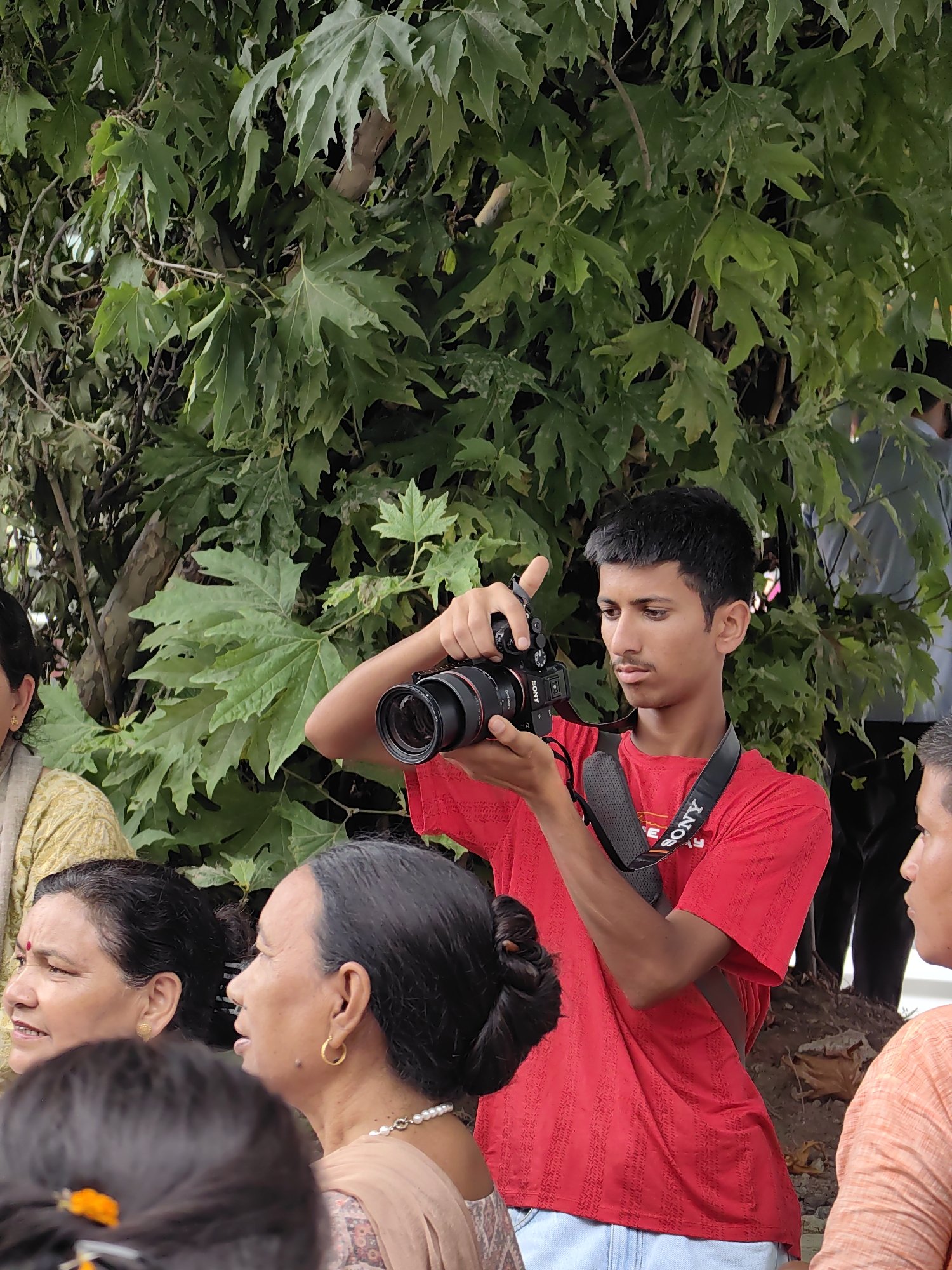A young man in a red t-shirt is holding a black camera with a prominent lens, looking through the viewfinder as if taking a photograph. He is wearing a black camera strap with "SONY" branding visible. He is positioned in the foreground, slightly to the right of center, against a backdrop of lush green foliage, suggesting an outdoor setting. In the middle ground, to the left of the frame, two women are visible in profile. The woman on the far left is wearing a yellow garment and has a red bindi on her forehead. Next to her, another woman wears a pearl necklace and gold hoop earrings. The background shows a blurred image of other people and more greenery, implying a public gathering or event in a park or natural area. The overall scene appears to be in Manali, India, given the presence of the Hindi word "SONY" on the strap and the natural scenery typical of the region.