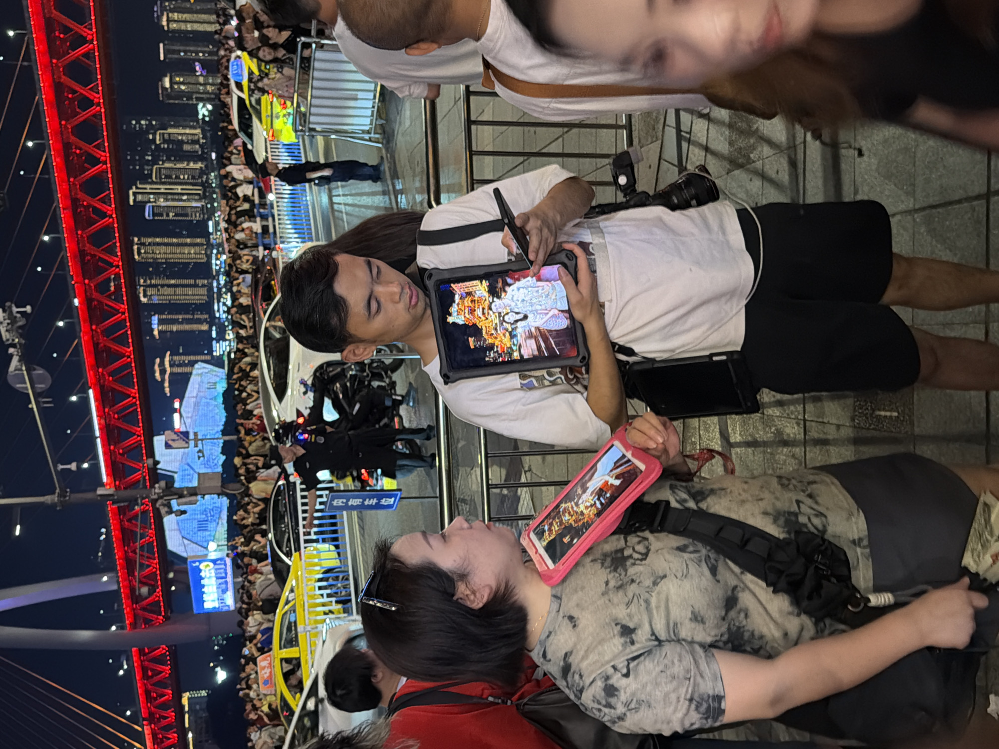 The image captures an outdoor night scene in Yuzhong District, China, likely during an event or gathering. In the foreground, a young man is engrossed in using a tablet with a stylus, suggesting he might be an artist or designer at work. The tablet displays an intricate illustration, possibly a traditional Chinese artwork. He wears a white t-shirt and black shorts.
Beside him, a young woman is looking at her tablet, which is encased in a bright pink cover. Her tablet screen also shows an image, possibly a photo or digital art. She is wearing a patterned t-shirt and appears engaged with her device.
In the background, a large, illuminated red bridge structure is visible, indicating an urban setting with significant infrastructure. A crowd of people is gathered, some standing and others seated, suggesting a public event such as a festival, concert, or viewing of a light show. Several vehicles, including what appear to be police cars or service vehicles, are present, hinting at crowd management or event security.
The setting is illuminated by artificial lights, including the vibrant red of the bridge and city lights in the distance, creating a lively nighttime atmosphere. The presence of numerous people and the festive lighting suggest a significant event is underway. The overall mood appears to be one of active engagement and public enjoyment.