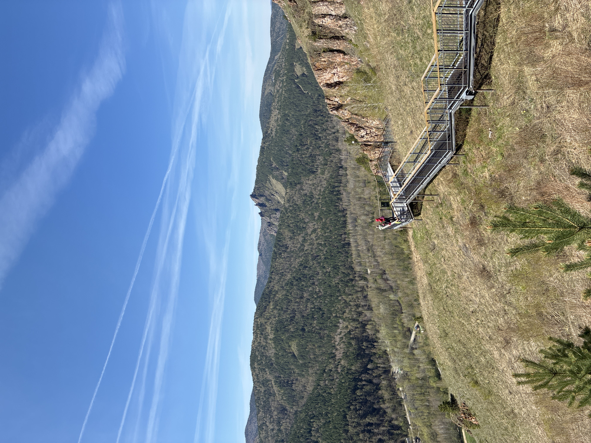 A landscape oriented outdoor shot showing a bright blue sky streaked with white contrails, a distant tree covered mountain range, a cliff face, a grassy slope, and a metal staircase descending the slope. There are a few small figures on the staircase, appearing to be people. The location is Unknown, Unknown. A few evergreen branches are visible in the bottom right corner.