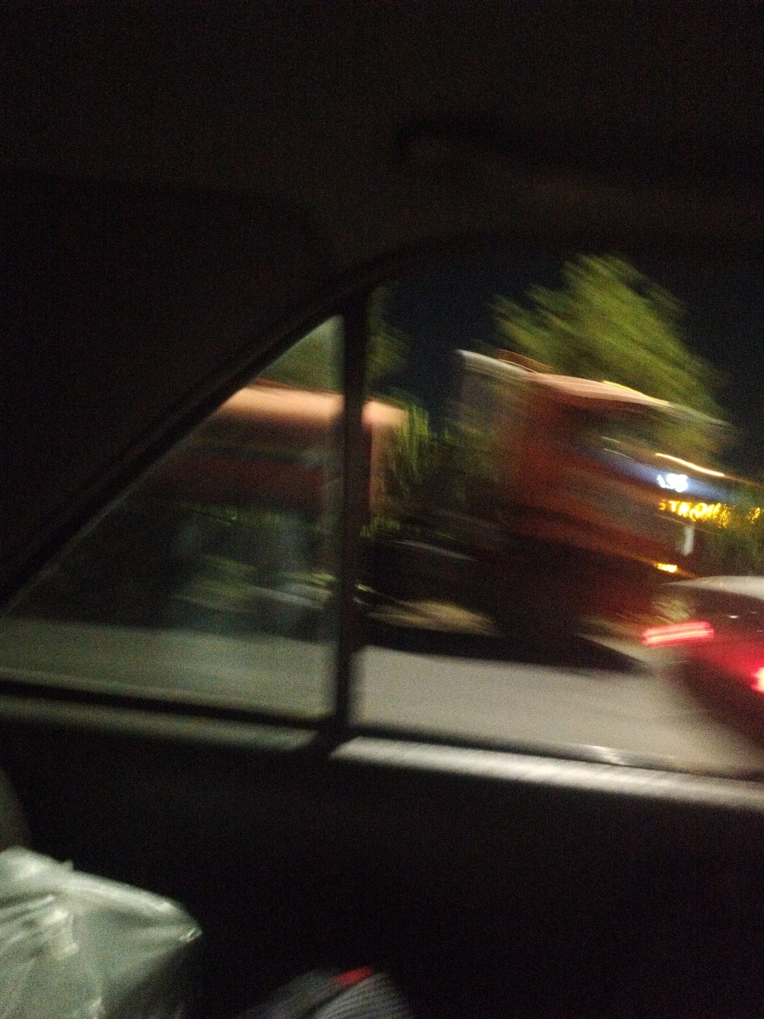 A motion-blurred, night-time view from inside a vehicle shows a street scene in Shymkent, Kazakhstan. The foreground is dominated by the interior of the vehicle, with a dark window frame on the left. Through the window, a large, out-of-focus vehicle, possibly a truck or bus, is visible. It is orange or red with some bright, blurred lights, suggesting it is in motion. The background features some greenery and illuminated buildings, but details are obscured by the low light and motion blur. There are no visible people in a recognizable state. The image suggests a drive through the city at night.