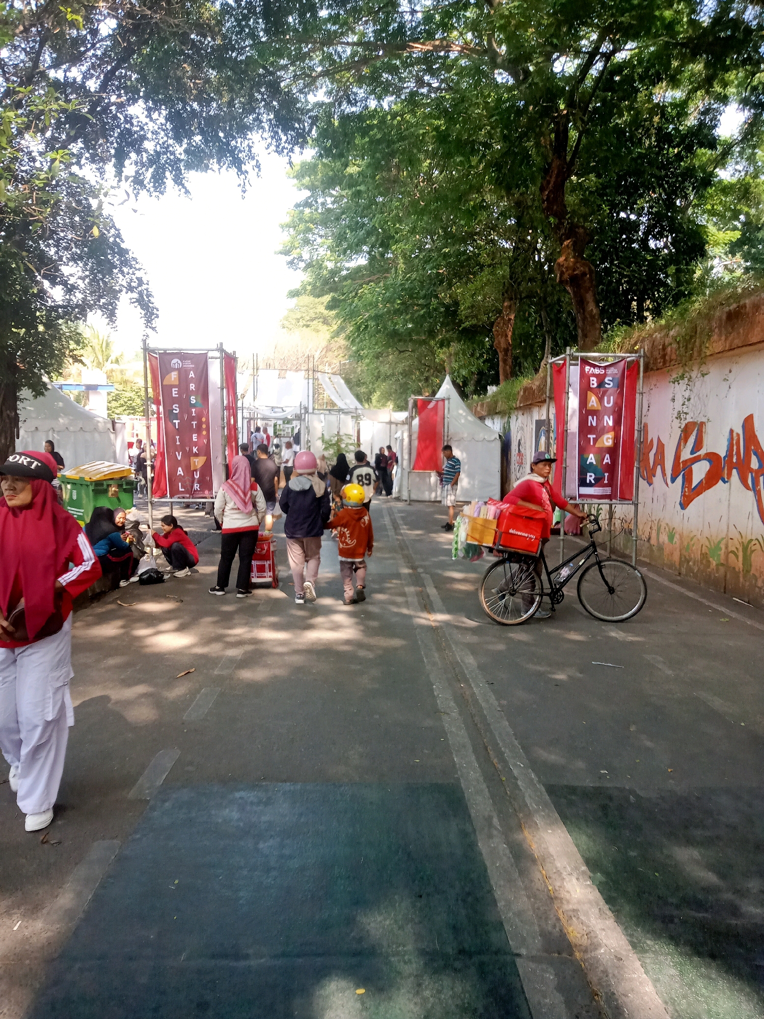 A sunny outdoor scene shows a street lined with tents and banners, suggesting a festival or market. The street is paved and has white and yellow lines marking lanes. Trees with lush green foliage overhang the scene, casting dappled shadows on the ground.
In the foreground, a woman wearing a red hijab and white pants stands facing left. Behind her, several people are gathered, some sitting on the ground and others standing. A green trash bin is visible to the left of the group.
Further down the street, a banner reads "FESTIVAL KOTA" in large letters. To the right, another banner, red with white text, displays "BS AU NT GA ARI". A person in a red shirt and a cap is pushing a bicycle with a large red cooler box attached to the back, loaded with colorful items, possibly for sale.
Several other people are walking in the middle ground, some wearing hijabs. Two children, one wearing a yellow helmet and the other a pink helmet, are also walking. In the background, more tents and structures are visible, and there are hints of buildings beyond. A graffiti-covered wall is on the far right of the image. The overall atmosphere appears to be lively and active. This scene is located in the city of East Jakarta, Indonesia.