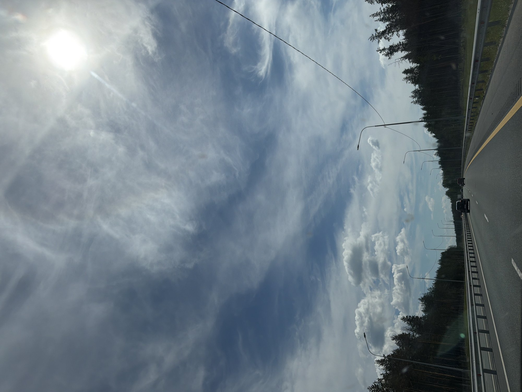 The image depicts a highway scene in the city of Velet'ma, Russia, under a partially cloudy sky. The sun is visible in the upper right corner, creating lens flare. A paved road with clear lane markings stretches into the distance, bordered by a metal guardrail. On the left side of the road, a dense forest of pine trees lines the highway. Several streetlights are spaced along the roadside. A few cars are visible on the road, traveling in the same direction. The overall environment suggests a daytime drive through a wooded area on a highway.