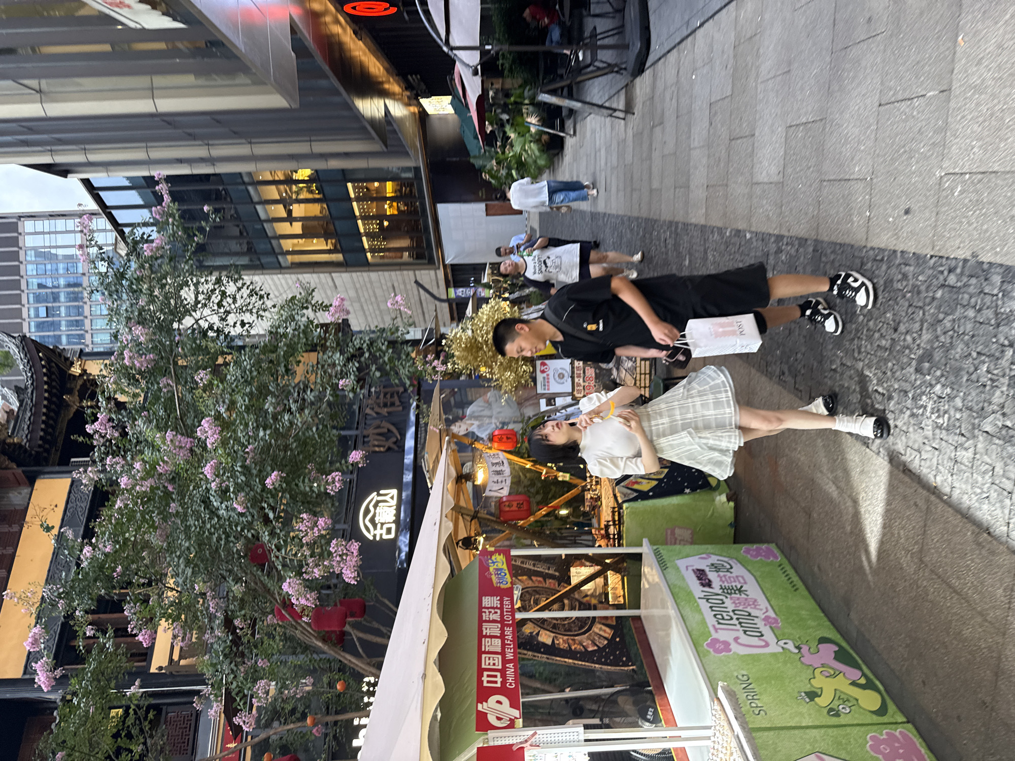This is a ground-level, medium shot image taken in Yuzhong District, China, depicting a street scene with a variety of elements. In the foreground, a young woman with short black hair, wearing a white puff-sleeved top and a light-colored plaid skirt, stands to the right of a vendor's stall. She is holding a yellow object and looking to the right. Adjacent to her, a young man in a black t-shirt and black shorts, wearing black and white sneakers, holds a white shopping bag in his left hand. He is looking towards the woman. The vendor's stall in the foreground is green and white, with a red sign that says "CHINA WELFARE LOTTERY" in English and Chinese characters. The stall also displays the text "SPRING Trendy Camp."
Behind the stall, pink flowers bloom on a tree. Traditional Chinese architecture with dark tiled roofs is visible in the background on the left. Further in the background, modern buildings with glass facades can be seen. People are walking in the background along the paved street, and there are tables and umbrellas set up, suggesting an outdoor dining or seating area. Red lanterns hang from the tree and the architecture. The street is paved with cobblestones and large rectangular tiles. The lighting suggests it is daytime, possibly late afternoon, with some shadows present.