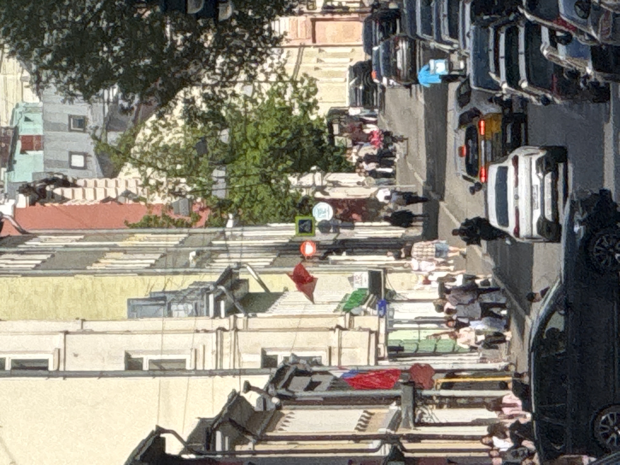 The image shows a street scene in Moscow, Russia, viewed from an elevated position. The camera's orientation is such that the streets and buildings appear to be rotated.
The architecture consists of buildings with light-colored facades, flat roofs, and some visible rooftops. Many buildings have air conditioning units and laundry hanging out on the balconies.
A street runs along the right side of the image. Several cars and trucks are parked closely together. There are pedestrians walking along the sidewalk.