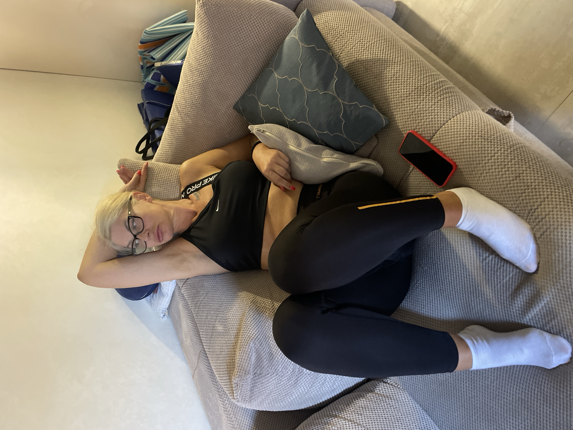 An adult woman with blonde hair and glasses is relaxing on a couch in an indoor setting, likely a living room. She is wearing a black sports bra, black leggings, and white ankle socks. Her right arm is raised and resting on her head, and she is looking to her left with a neutral expression. A red smartphone is placed on the couch to her right. Behind her, on the couch, are several pillows, and further back, there are bags that appear to be travel bags. The lighting suggests it is daytime. The scene is presumed to be in Budva, Montenegro, based on the provided context, but the visual elements do not specifically confirm this location.