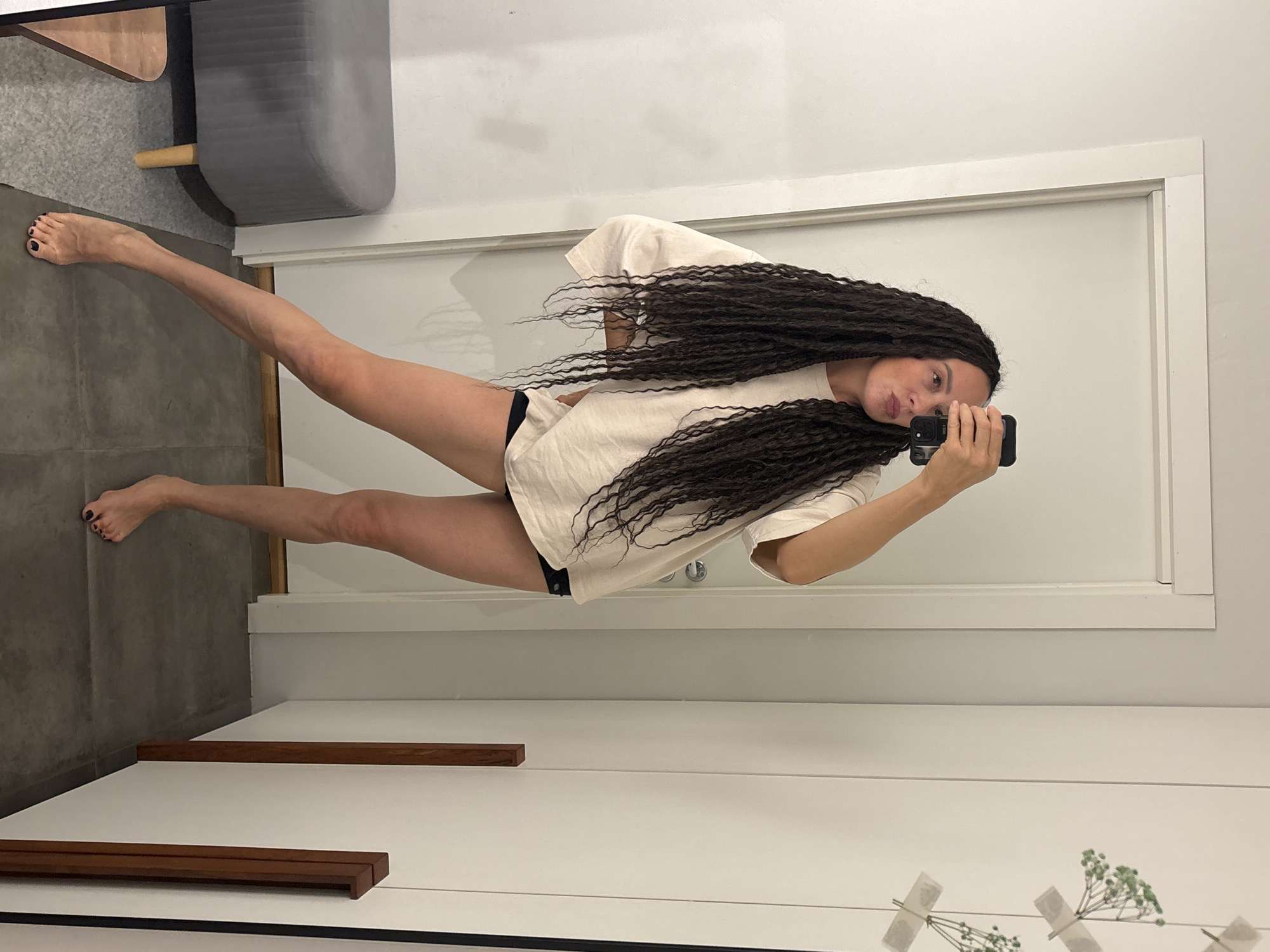 This image captures an adult female posing for a mirror selfie within an indoor setting in the city of Chaikovsky, Russia. The woman has very long, dark, dreadlock-style hair that extends past her waist. She is wearing a loose-fitting, light beige or off-white long-sleeve t-shirt and dark, possibly black, underwear or shorts. She is barefoot, with dark nail polish visible on her toenails. Her left hand holds a black smartphone, likely an iPhone, capturing her reflection, while her right arm is bent with her hand on her hip. She is leaning slightly to her left, with her left leg bent and her right leg extended, adopting a casual pose.
The background features a white door with a light-colored frame on the left, and a white wall with two vertical dark wood accent strips on the right. Between these strips, small sprigs of green foliage are taped to the wall, serving as a minimalist decorative element. In the lower-left corner, a gray upholstered stool with light wooden legs is visible. The floor transitions from a dark gray tiled area in the foreground to a lighter gray carpeted section under the stool. The scene is well-lit, conveying a clean and modern aesthetic.
