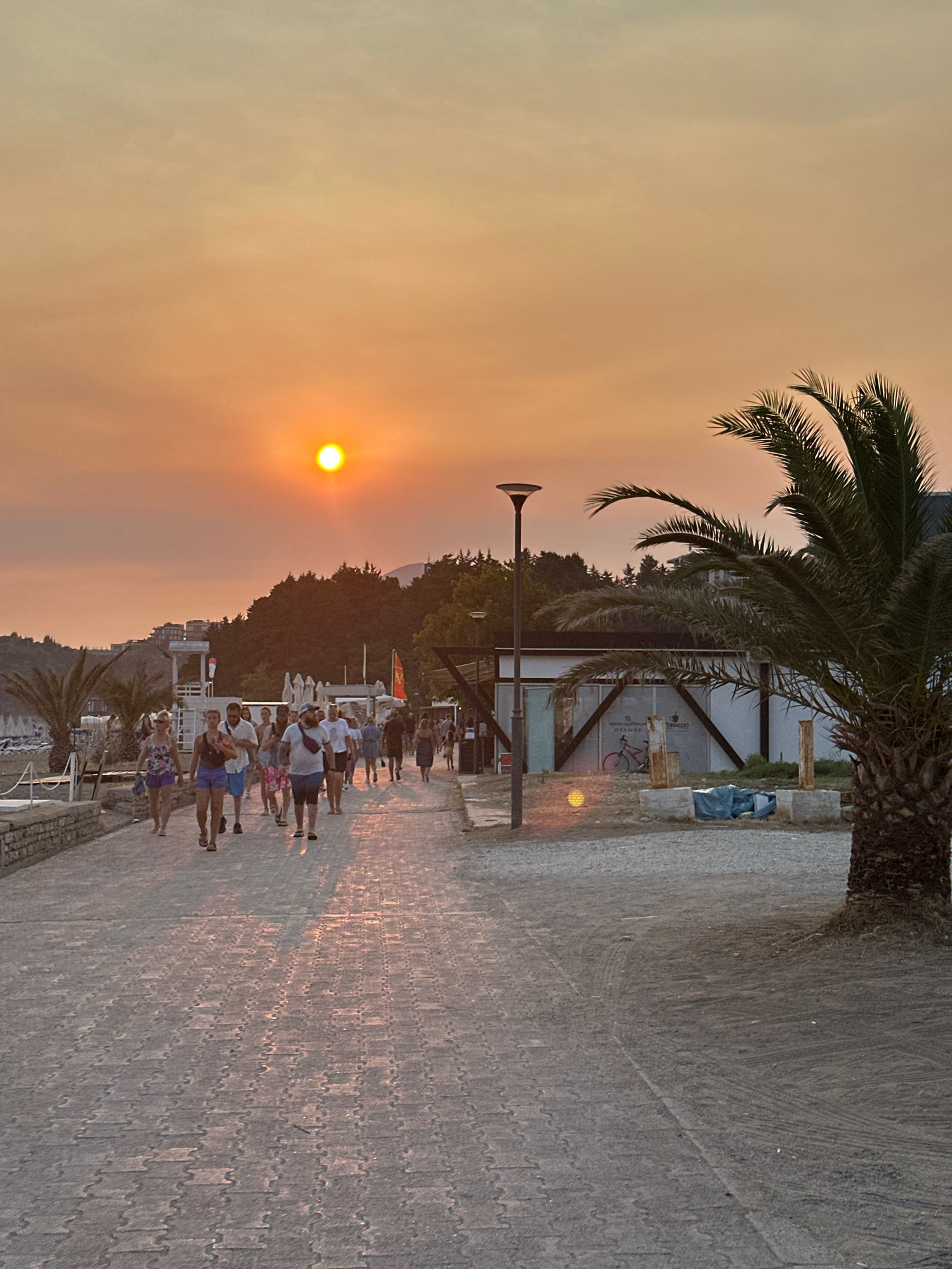 The image depicts a sunset scene on a paved walkway in Boreti, Montenegro. The sun, a bright orange orb, is visible in the hazy, orange-toned sky. A group of people are walking along the path, casting long shadows. The walkway is made of interlocking grey paving stones. To the right, a palm tree stands prominently, with a modern-looking building partially visible behind it. The building appears to be a small structure with a glass facade and a wooden frame. There is a lamppost next to the building. In the background, trees and some buildings can be seen on a hillside. The overall atmosphere is warm and serene, typical of a coastal town at dusk.
