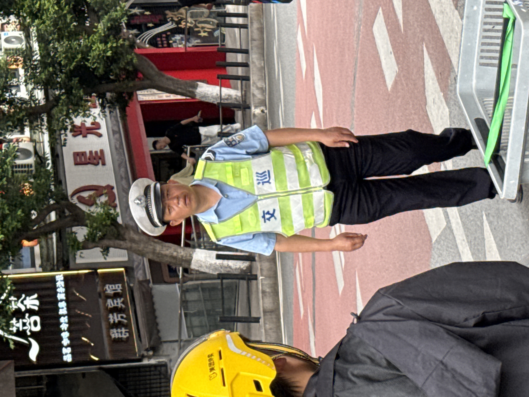 A photograph shows a man in a light blue short-sleeved shirt, black pants, and a white hat, over which he is wearing a yellow reflective vest with Chinese characters that appear to identify him as traffic police. The man is facing forward and has his arms at his sides. He is standing on a sidewalk or road marked with white and red lines.
To his right and slightly below is part of another person wearing a yellow helmet and a black coat or jacket. Behind the traffic police man is the entrance to a building and signage in Chinese. The location appears to be an urban area, plausibly the city of Liangjiang, China.