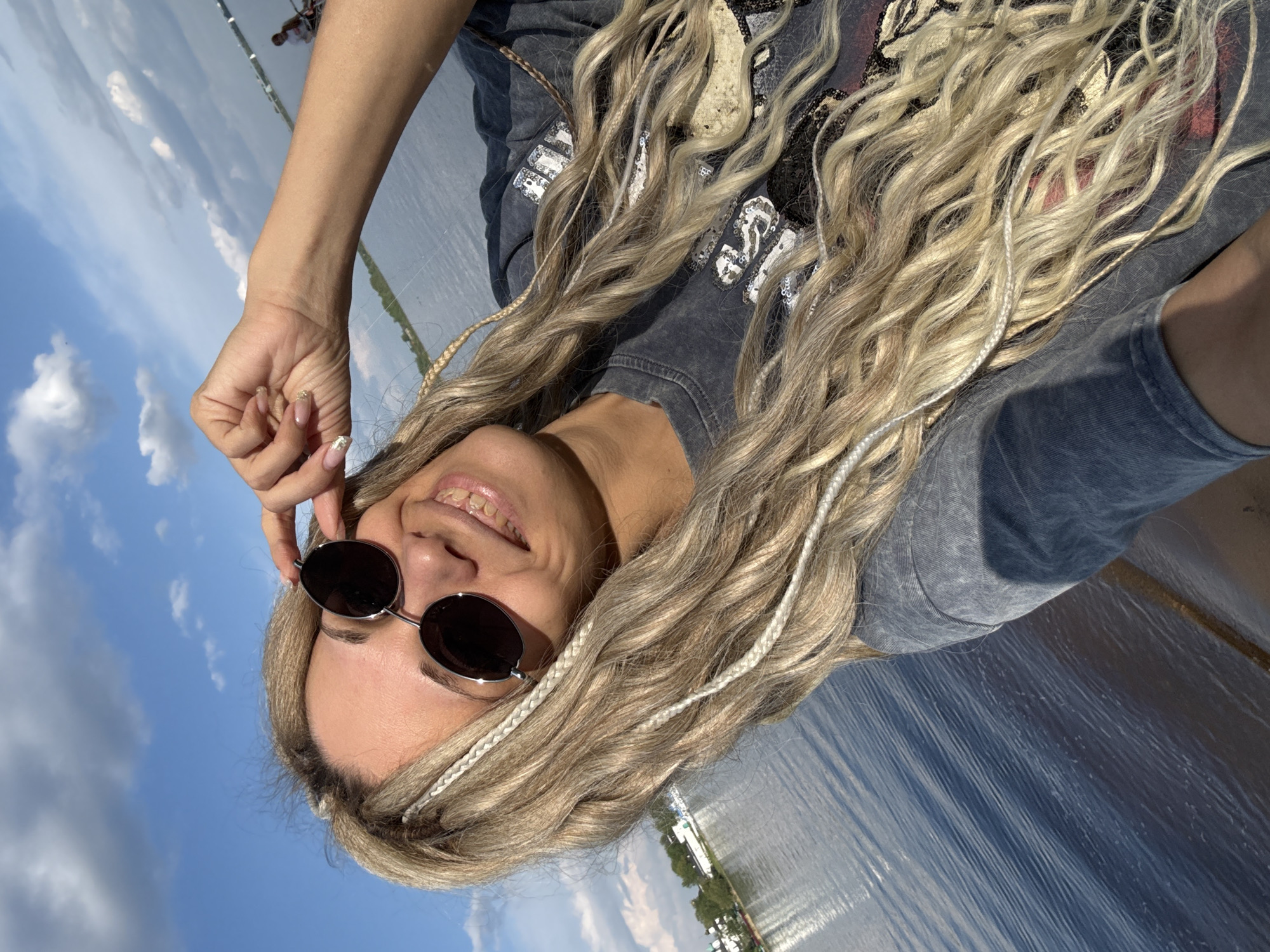 A low-angle outdoor selfie shows a woman with light-colored, textured, and braided hair wearing round sunglasses and a printed gray t-shirt. She is smiling. The background includes a body of water and a blue sky with clouds. The location is in the city of Rusanovo, Russia.