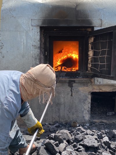A man wearing a light-colored shirt, gloves, and a tan head covering stands outside of a metal furnace in Kandahar, Afghanistan. The furnace door is open, revealing intense flames and a shovel inside. A pile of coal sits in front of the furnace. The surrounding environment appears industrial and gritty.