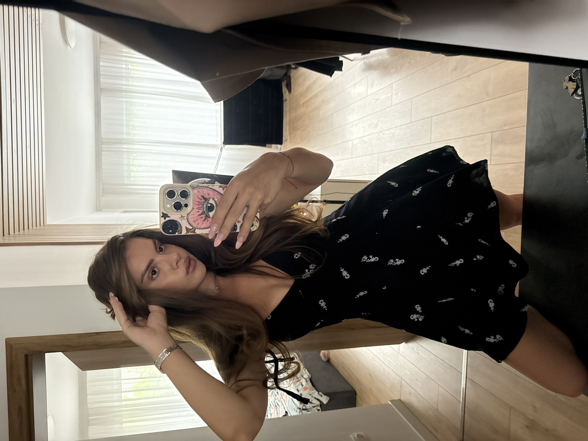 A woman is taking a mirror selfie in a room. She is wearing a short black dress with a floral pattern and a V-neck. She has long brown hair and is wearing a silver bracelet on her left wrist. In her right hand, she is holding a smartphone with a decorative case. She is standing in front of a full-length mirror, which reflects the room. The room appears to be a bedroom or dressing area, with a bed visible in the background to the left and light-colored curtains covering a window. The flooring is light-colored wood.