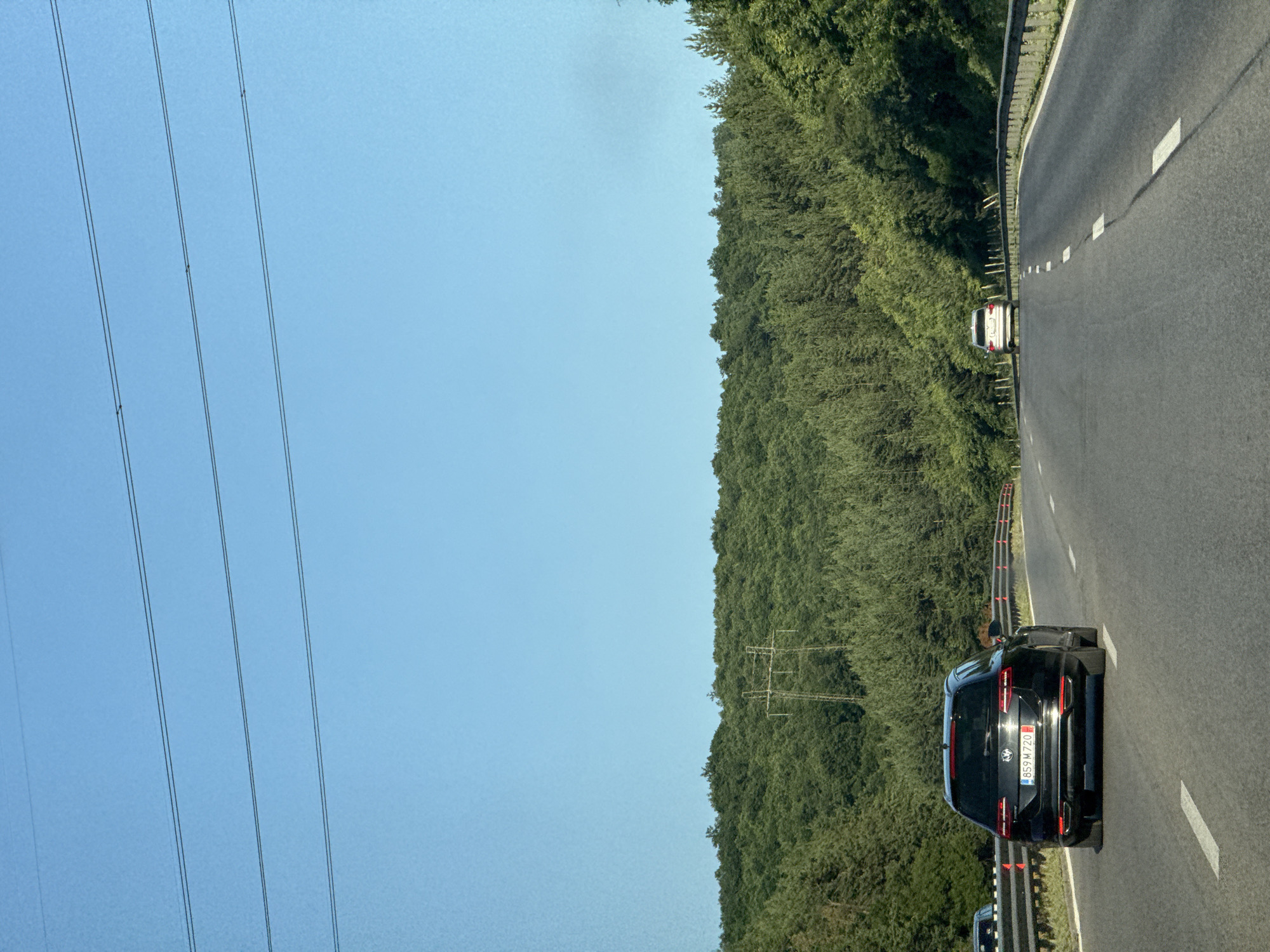 The image depicts a highway scene in Eleshnitsa, Bulgaria, with a clear blue sky and lush green trees lining the road. The perspective is from inside a vehicle, looking forward and slightly to the right. In the foreground, a black SUV is driving on the right lane of the highway. Its license plate is visible and reads "B59 M 720". Further ahead, a white car is visible in the distance. The highway is marked with white lines, and a metal guardrail separates the road from the densely wooded hillside. Overhead power lines are also visible on the left side of the image. The overall impression is that of a sunny day, likely mid-day, with good visibility for driving. The scene conveys a sense of travel and movement.