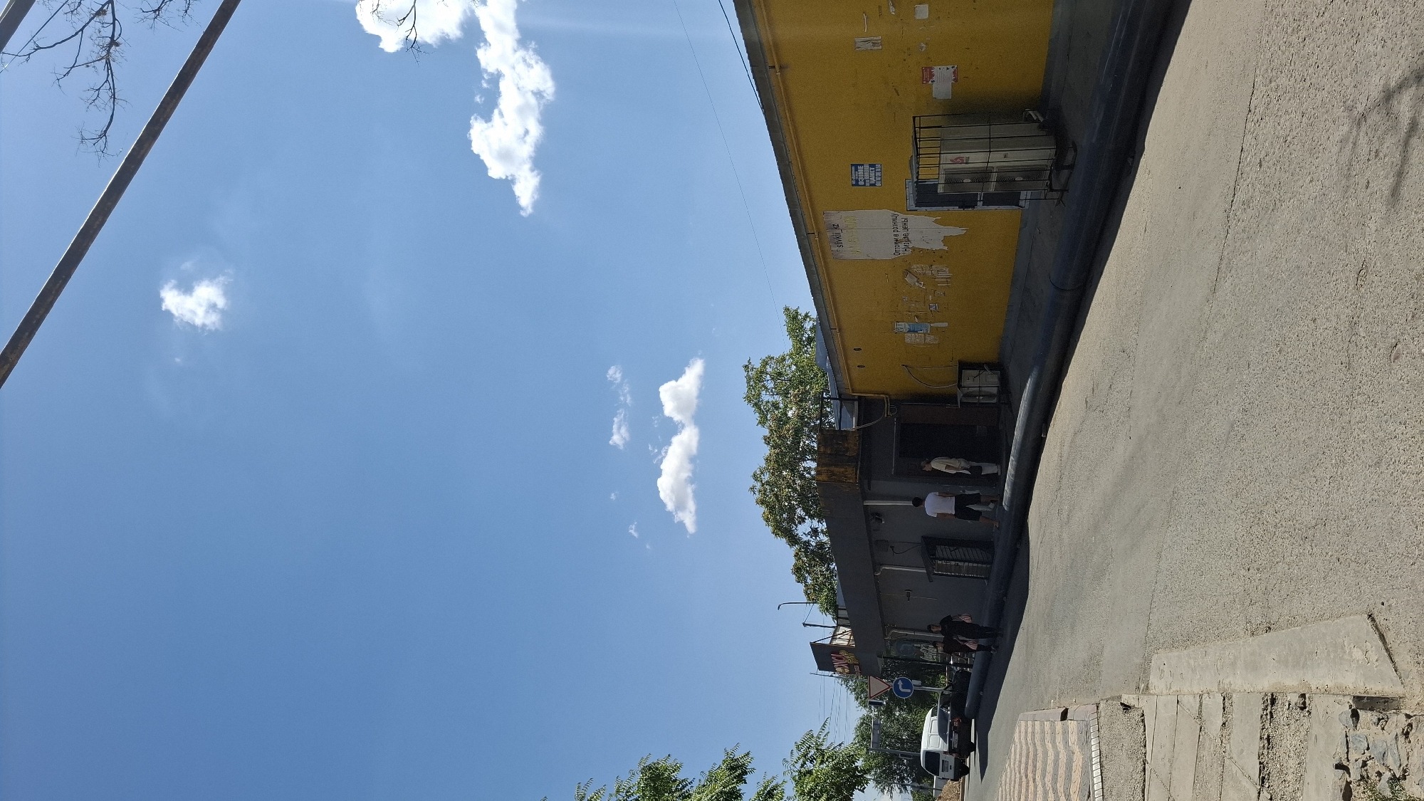 This is a street-level, vertical, eye-level, outdoor, daytime, full shot image taken in Shymkent, Kazakhstan. The scene depicts a street with a paved road and a concrete sidewalk on the left. A yellow building with a gray facade stands to the right, featuring several posters and flyers on its yellow wall. In front of the building, there is an entrance with a dark opening, and two individuals are standing near it. One person is wearing a white t-shirt and shorts, while the other is wearing a light-colored shirt and pants. Further down the street, a white van and a black car are parked. The sky above is blue with scattered white clouds. The overall impression is of a sunny day in a somewhat urban or commercial area.
