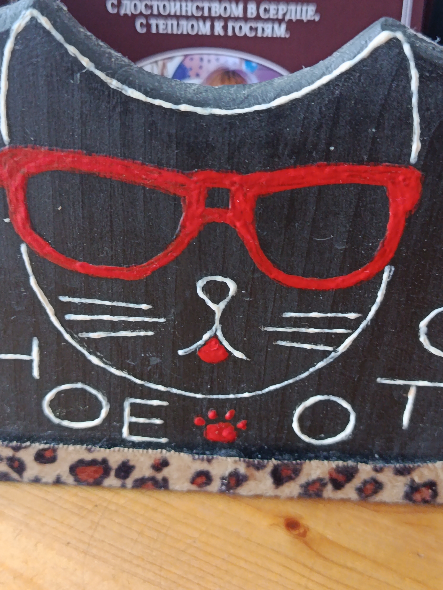 The image is a close-up, vertical shot of a black, wooden cat-shaped object, possibly a decorative item or a holder. The cat is adorned with bright red, oversized sunglasses, and its face is outlined in white paint, featuring whiskers, a small red nose, and a subtle smile. The white paint is also used to write what appears to be text on the lower part of the cat's face. The bottom edge of the object has a band of fabric with a leopard print pattern. The object is placed on a wooden surface, likely a table or shelf, and the lighting suggests it is indoors during the daytime. The overall mood is playful and whimsical due to the cartoonish design and bright colors.
Above the cat's head, there is some visible text in white Cyrillic lettering against a dark background, possibly part of a larger sign or label. The text reads: "С ДОСТОИНСТВОМ В СЕРДЦЕ, С ТЕПЛОМ К ГОСТЯМ." This translates to "With dignity in heart, with warmth to guests."
Given the location context of Braslaw, Belarus, the text implies a sentiment of hospitality or a welcoming atmosphere. The object itself, with its cat motif and playful glasses, could be a souvenir, a decorative piece for a home, or a shop item. The close-up framing emphasizes the details of the cat's face and the applied decorations.