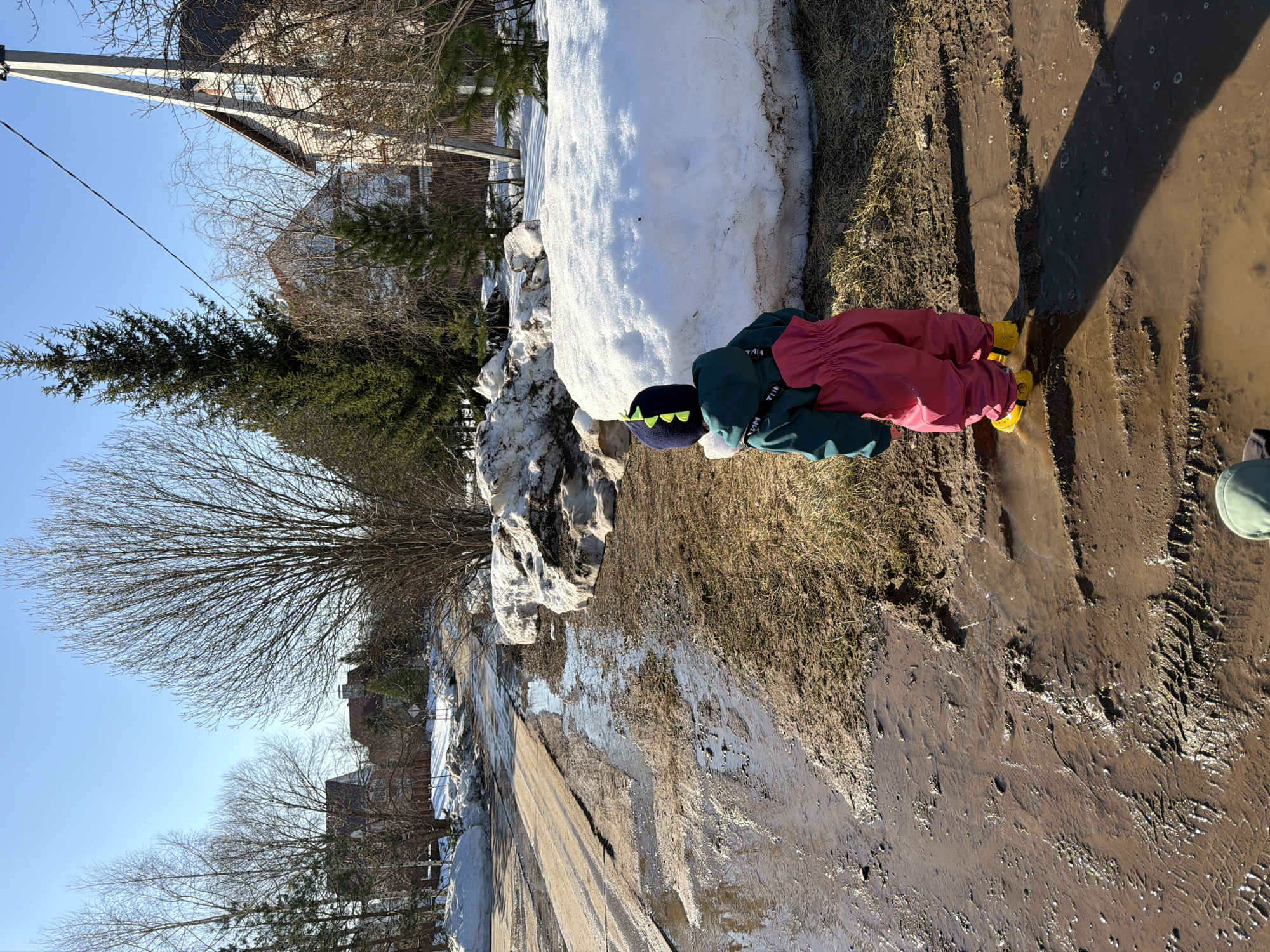 The image shows a child playing in a muddy area near a snowdrift. The child is wearing a green jacket, pink pants, and yellow boots, as well as a dark blue hat with spikes. The child is crouched down, facing the mud. The background includes a snowy area, buildings, and trees. The location is in the city of Chaikovsky, Russia.