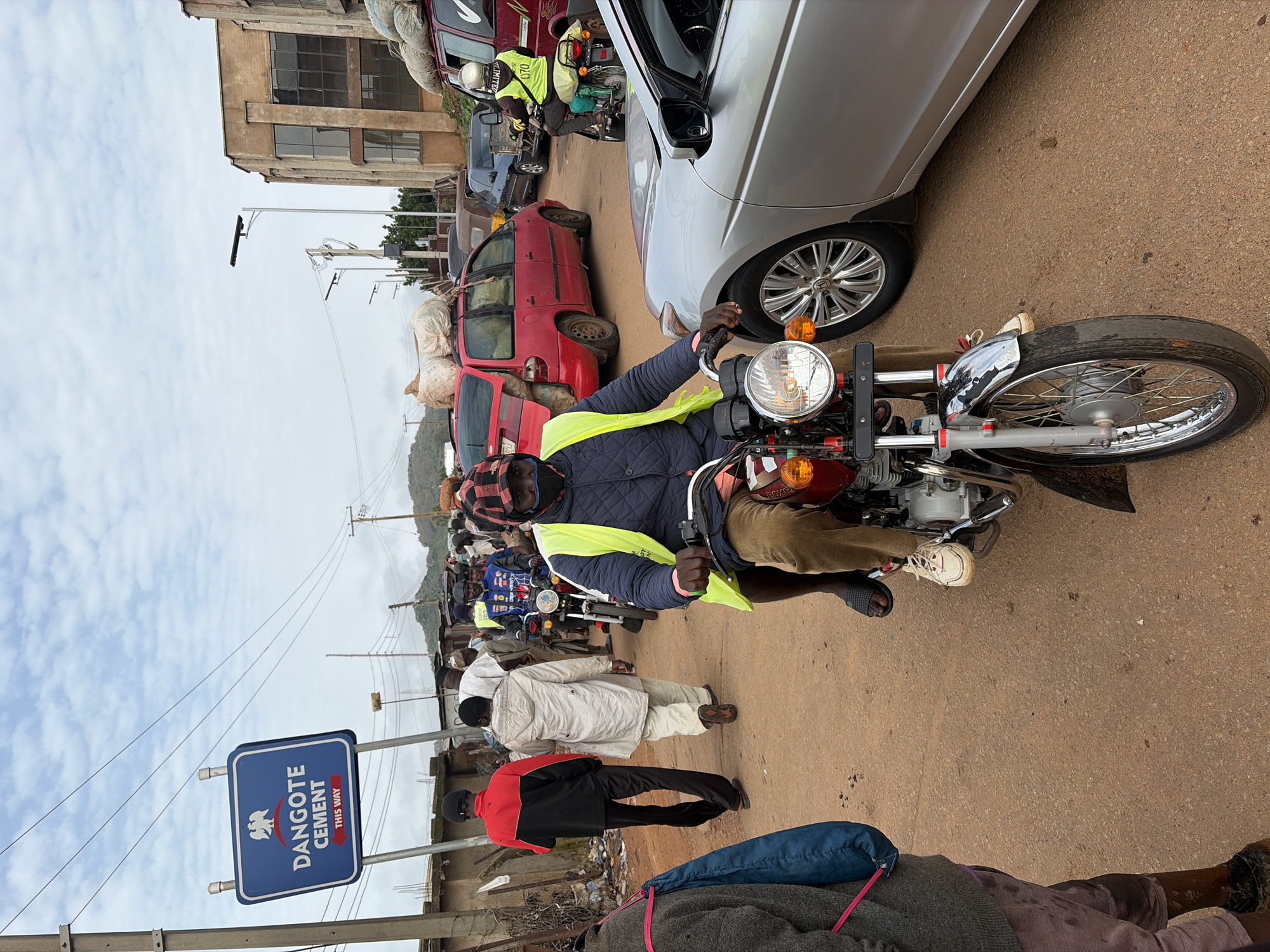 A man wearing a reflective yellow vest and a checkered bandana over his face rides a red motorcycle through a crowded street. He is on the right side of the image, and the motorcycle is facing towards the viewer. In the foreground, the motorcycle's front wheel, headlight, and handlebars are visible. The man's legs are clad in brown trousers, and he wears white sneakers and black flip-flops. Behind him, a silver car is parked on the right. To the left of the motorcycle, other people are walking and riding motorcycles. In the background, red and blue cars are present, along with buildings and a sign that reads "DANGOTE CEMENT THIS WAY." The ground is dirt or gravel. The sky is overcast. The location is Gura Top, Nigeria.