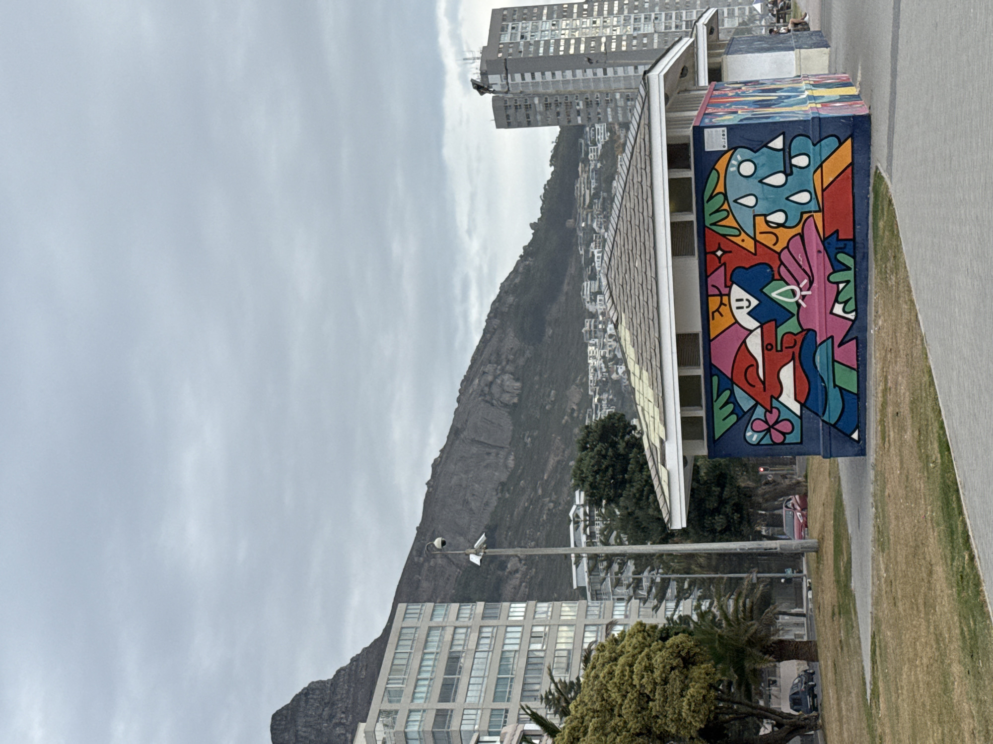 The image shows a scene in Cape Town, South Africa, with Table Mountain visible in the background under a cloudy sky. In the foreground, there is a building with a mural painted on its side, featuring colorful, abstract figures and shapes. A grassy area borders a paved walkway next to the building. The middle ground includes trees, street lighting and buildings, including a high-rise. Several more buildings are nestled on the lower slopes of Table Mountain.