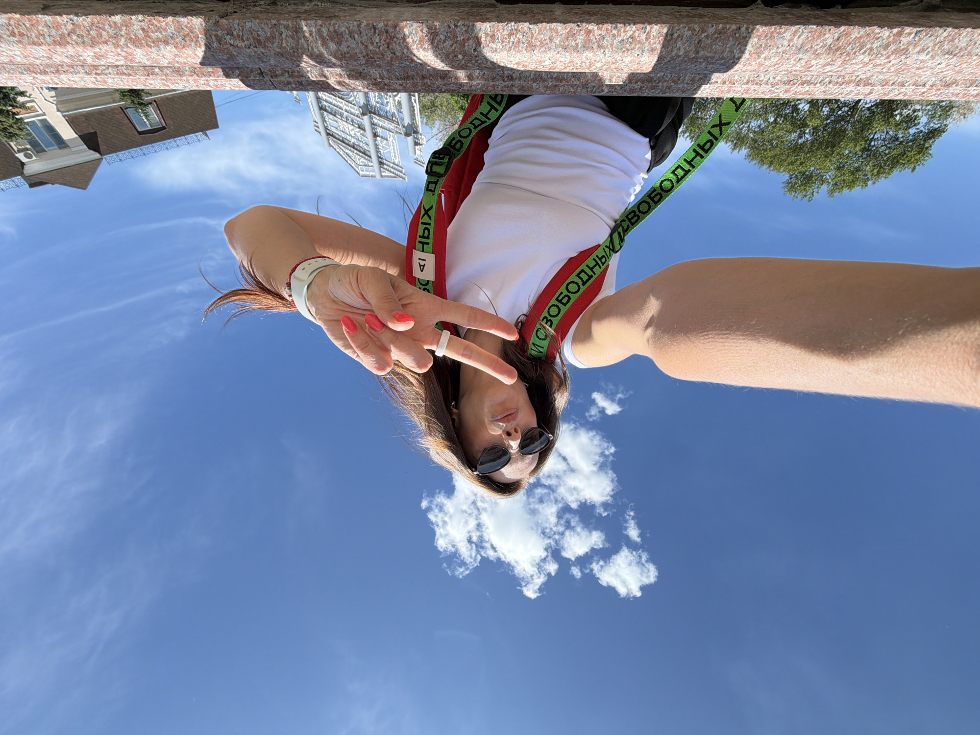 A woman is taking a selfie outdoors on a sunny day in Birobidzhan, Russia. She is wearing sunglasses, a white tank top, and black pants, with a red strap from a bag or backpack visible across her chest. She is making a peace sign with her right hand and has bright red nail polish. The background is a bright blue sky with some white clouds. In the foreground, there is a textured, pinkish-red surface, possibly a bench or a wall. Behind the woman, there are some trees and buildings, including a modern-looking structure with a large billboard. The overall scene suggests a casual, outdoor activity.
