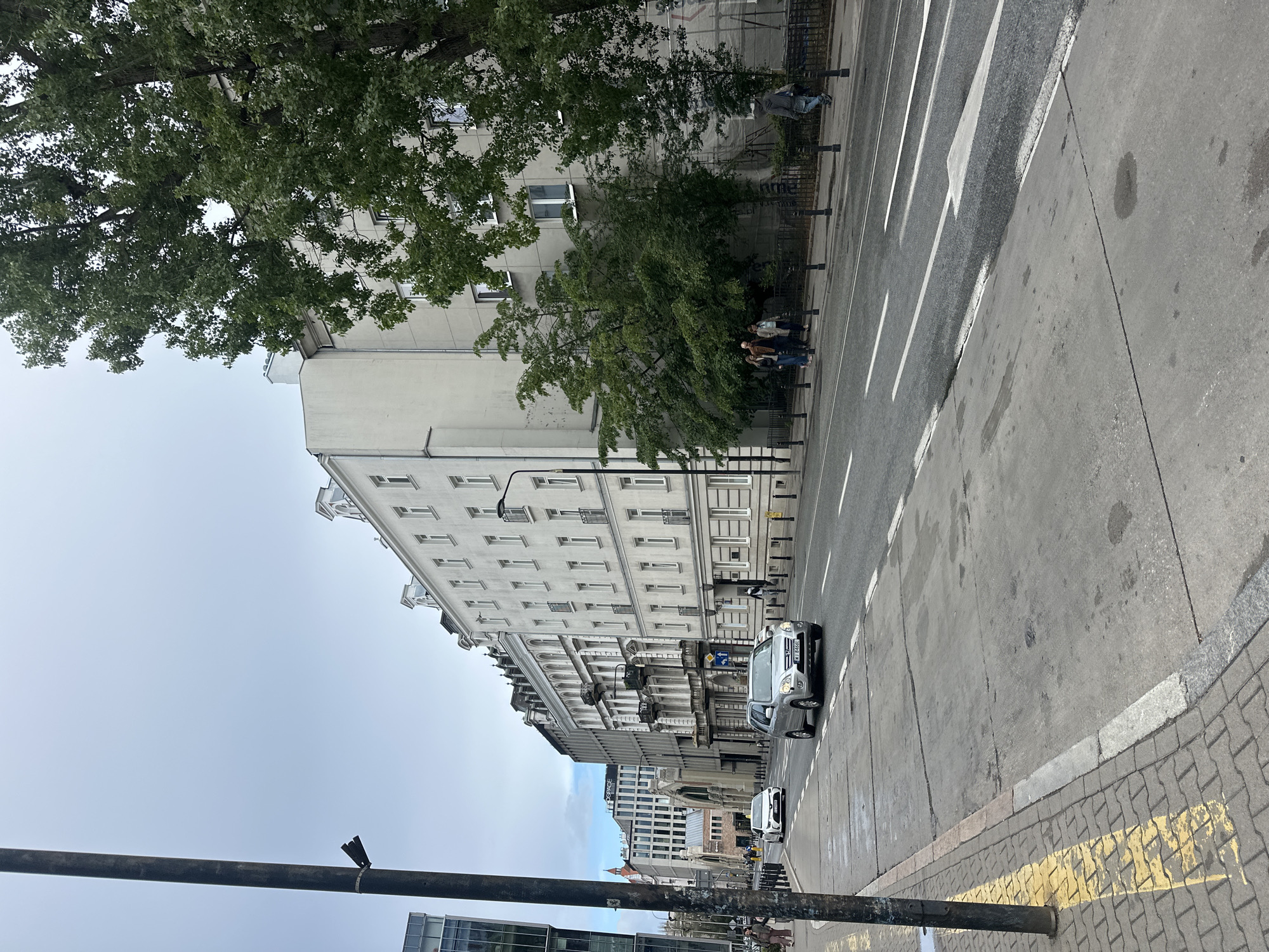 The image depicts a street scene in Warsaw, Poland, specifically on Aleje Jerozolimskie. In the foreground, a gray sidewalk with yellow markings runs alongside the street. A dark metal lamppost stands on the left side. The street itself is paved and features white lane markings.
Several vehicles are present on the road. A silver SUV is in the right lane, and a white sedan is in the left lane. In the background, a row of apartment buildings with white facades and ornate architectural details lines the street. Some of these buildings have balconies and dormer windows. Tall trees with green foliage are interspersed along the sidewalk and in front of the buildings, partially obscuring the facades.
A few pedestrians are visible. On the sidewalk, a group of three individuals walks away from the camera. Further down the street, another person walks towards the camera on the right side of the road. A person in a gray hooded jacket is seen walking on the sidewalk on the right side of the image, moving away from the camera.
In the background, more modern buildings are visible, including one with the text "OSSPACE" on it. The sky is overcast and a pale blue. The overall impression is a city street on a cloudy day, with a mix of historic and contemporary architecture.