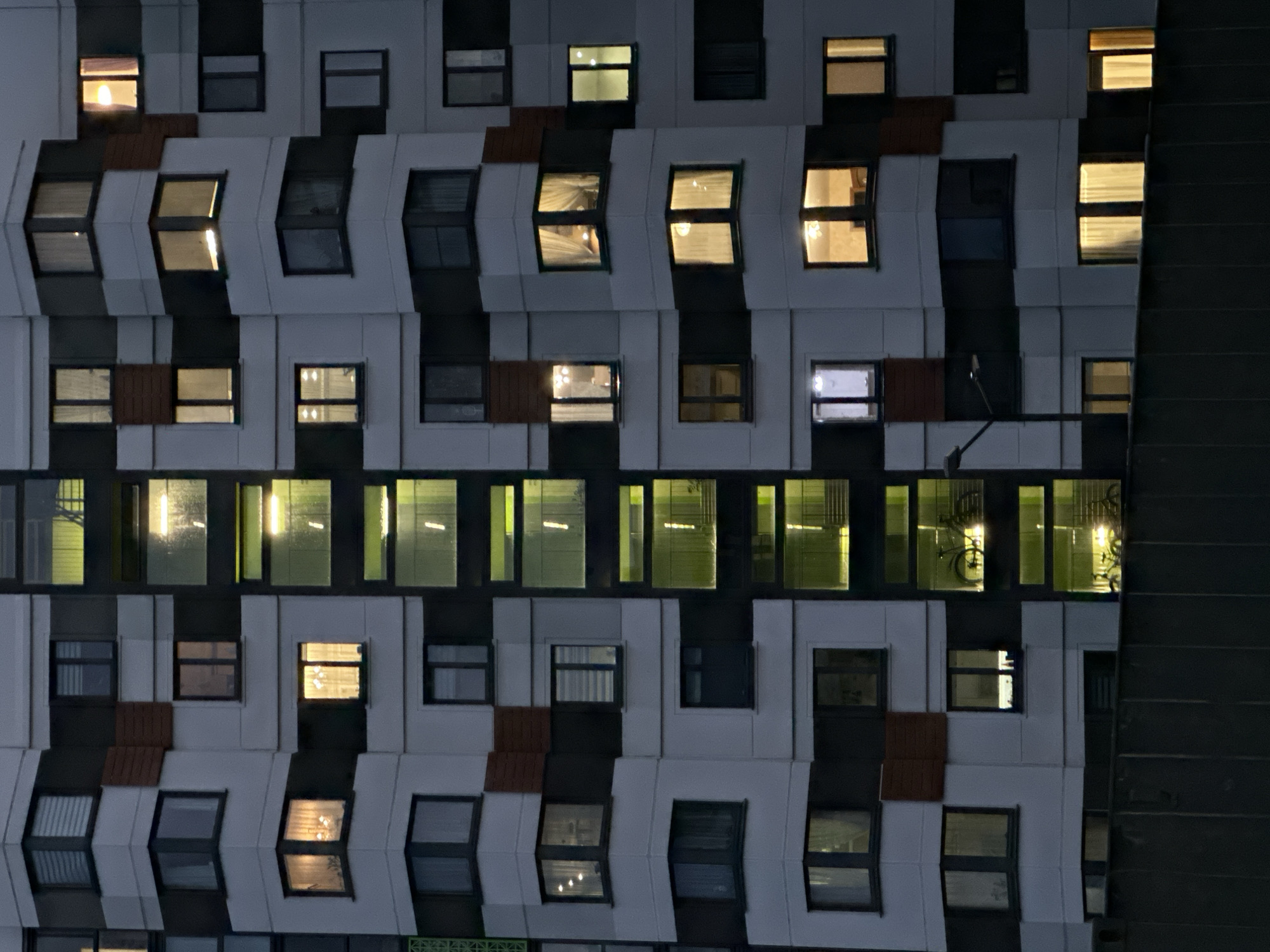 The image depicts the facade of a building in Бараўлянскі сельскі Савет, Belarus, at night. The building has a distinct architectural style with a pattern of white and dark grey or black sections. The windows are mostly rectangular, and some are lit, revealing interior scenes. Some windows reflect indoor lighting. One window appears to have a bicycle hanging inside. A streetlight or pole is visible near the right edge of the building, and the adjacent building is very dark. The overall scene suggests an urban residential environment.