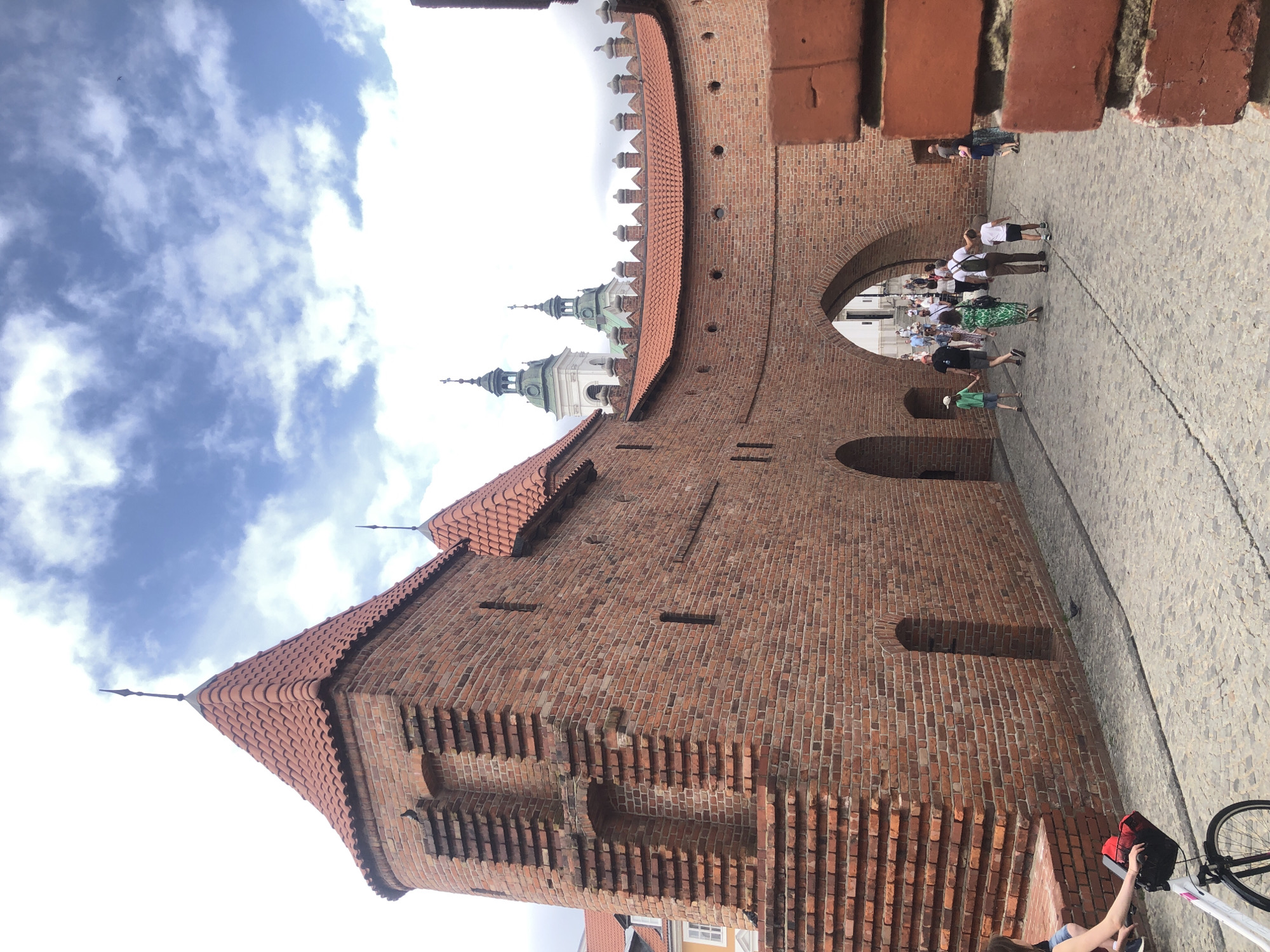 The image shows a view of the Barbican in Warsaw, Poland. The Barbican is a medieval fortification that is part of the Old Town's defensive walls. In the foreground, a brick wall and a portion of a tower are visible. The tower has a red tiled roof with spires. The wall leads to an arched gateway through which people are walking. The ground is paved with cobblestones. A woman is visible on the left side of the image, pushing a bicycle with a red basket on the handlebars. In the background, two church towers with green roofs are visible above the city walls. The sky is partly cloudy. The scene depicts a historical site within a city, with a mix of ancient architecture and modern-day activity.