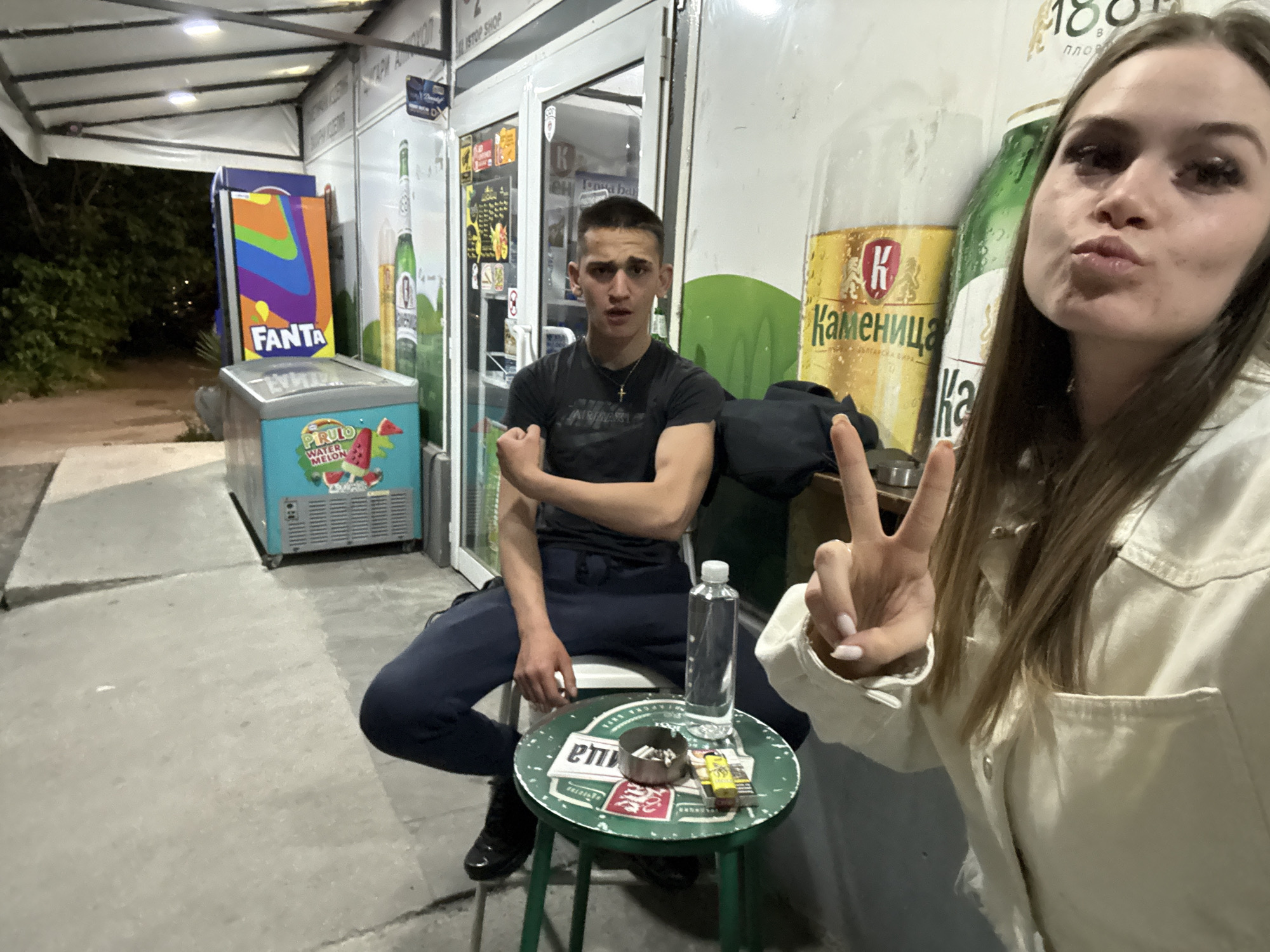 The image captures two individuals outside a shop in Plovdiv, Bulgaria. A young man sits on a green stool next to the shop's entrance, while a woman stands closer to the camera, partially obscuring the view with a peace sign. The man is dressed in dark clothing and is seated near a stool cluttered with an ashtray, cigarettes, and a water bottle. Behind him, the shop's exterior is visible, displaying advertisements for "FANTA" and local beer brands like "Каменица". A cooler labeled "PIRULO" is also nearby. The environment suggests an evening setting with street lighting. The overall impression is a candid snapshot of everyday life in the city.