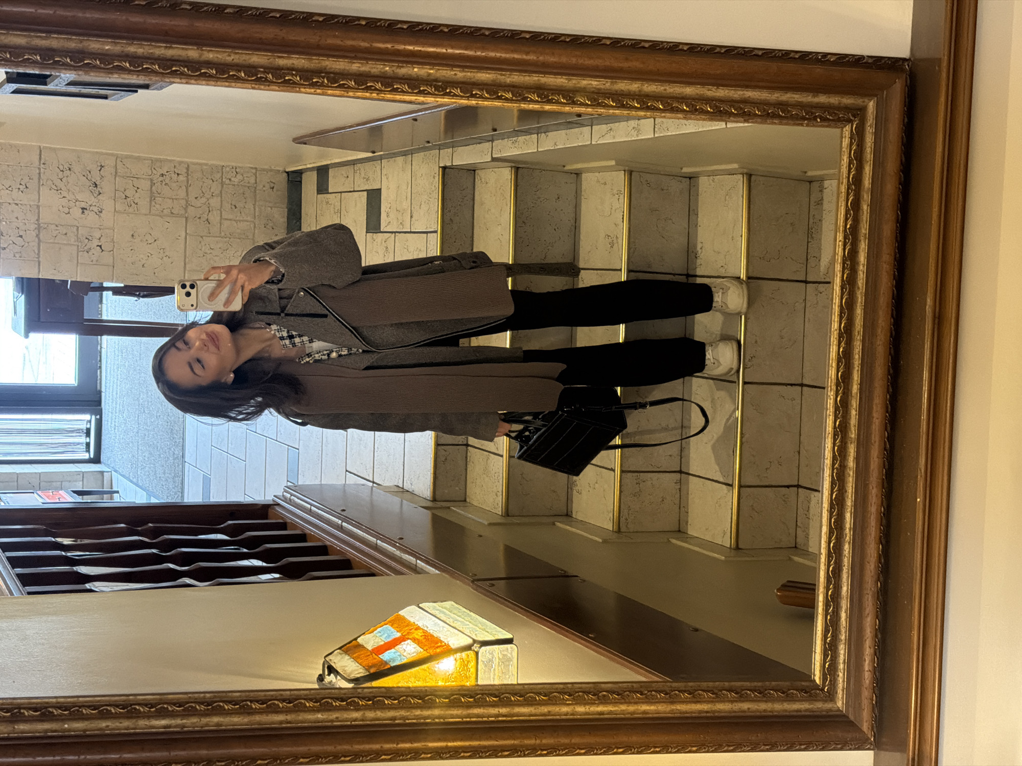 The image shows the reflection in a gold-framed mirror. The reflection shows a woman taking a selfie with her cell phone. She has long dark hair and is wearing a gray coat, a plaid top, black pants, and white sneakers. She is also carrying a black purse. The background of the reflection is a staircase with beige and gold tiles. The location appears to be an indoor staircase in the city of Krasnoyarsk, Russia. The frame of the mirror is visible, and a stained-glass lamp is situated beneath the mirror.
