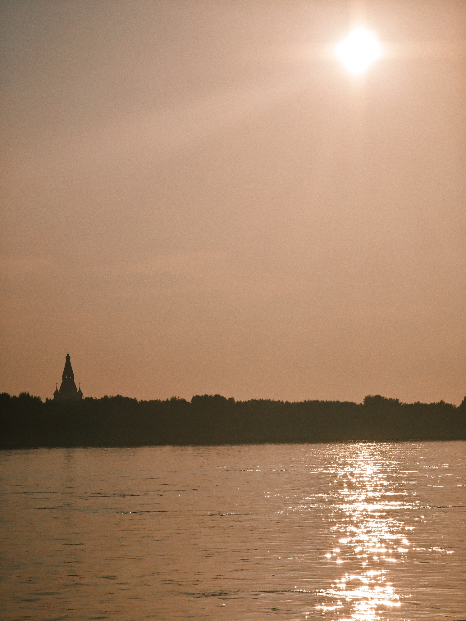 This media file depicts a serene landscape scene with a body of water in the foreground and a silhouetted tree line and a building in the background. The sun is bright and high in the sky, casting a warm, golden glow over the entire scene. The water reflects the sunlight, creating a shimmering effect.
The subjects in the scene are the natural elements: the water, the trees, and the sky. There are no people visible. The building appears to be a church with a tall spire and a cross on top, silhouetted against the sky. The tree line forms a dark band across the middle ground, suggesting a forest or dense vegetation.
The activity taking place is simply the passive observation of a natural landscape. The time of day is clearly daytime, given the bright sun. The weather appears clear and sunny. The location cues, such as the presence of a church and a dense tree line, along with the provided location context of Lesosibirsk, Russia, suggest a possibly rural or semi-rural setting in that region.
Notable details include the intense brightness of the sun, which creates a lens flare effect at the top of the image. The water's surface is textured with ripples, and the sunlight creates a dazzling reflection across it. The overall mood conveyed by the image is one of tranquility and natural beauty, enhanced by the warm, atmospheric lighting. There is no visible text in the image.