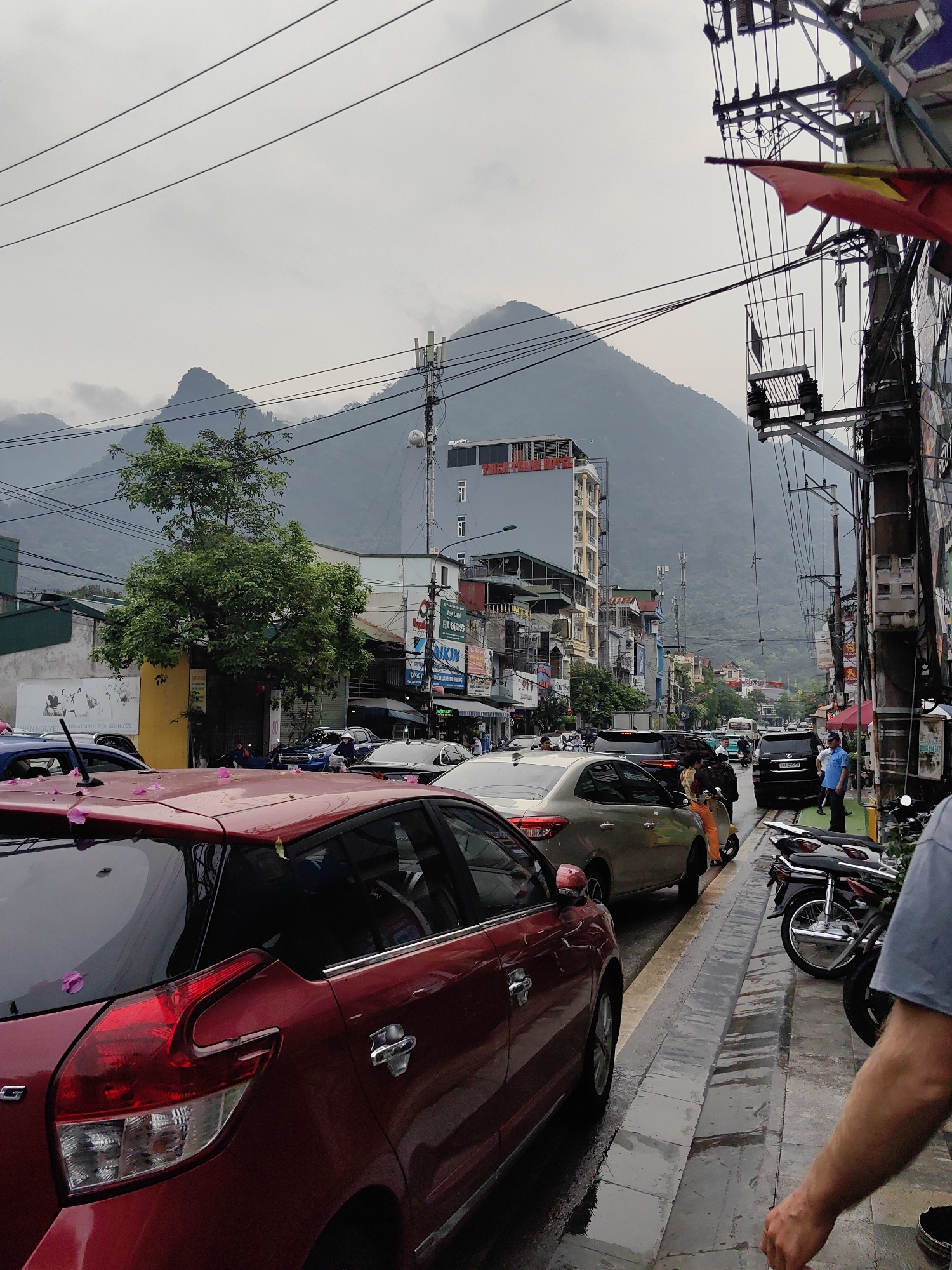 The image captures a street scene in Thành phố Hà Giang, Vietnam, characterized by a cloudy sky and a mountain backdrop. The street is lined with multi-story buildings and dense utility lines overhead. A traffic jam is visible, with cars of various colors, including a red hatchback in the foreground and a tan sedan next to it, filling the roadway. Two motorcycles are parked on the sidewalk. A man in a blue shirt stands near the sidewalk. Pink flower petals are scattered on the red car's surface.