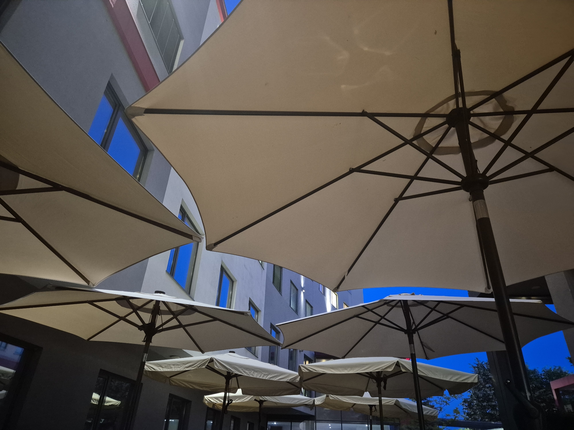 A low-angle, upward-looking photograph captures an outdoor seating area, likely at dusk or night, dominated by several large patio umbrellas. The umbrellas are uniformly beige or off-white with dark metal frames and spokes clearly visible from underneath, creating a patterned structure against the sky and building. Their angular shapes fill much of the foreground and midground, framing the background elements.
Behind and between the umbrellas stands a modern building with light grey or off-white walls. Multiple rectangular windows are visible on the building's facade, glowing with a striking, vivid blue light, which could be interior illumination or reflections of the twilight sky. Subtle hints of red architectural trim can be seen on parts of the building's exterior.
The sky, visible in gaps between the umbrellas and above the building, is a deep, clear blue, indicative of either dusk or early night. There are no clouds present. The overall lighting is dim, emphasizing the glow from the windows and the subtle illumination of the umbrella undersides.
The scene is devoid of people or discernible activity, conveying a calm and static atmosphere. The composition emphasizes geometric forms and the interplay of neutral tones with the vibrant blue light. The setting suggests a communal outdoor space, such as a hotel courtyard, a restaurant patio, or a similar urban leisure area. No visible text is present in the image.