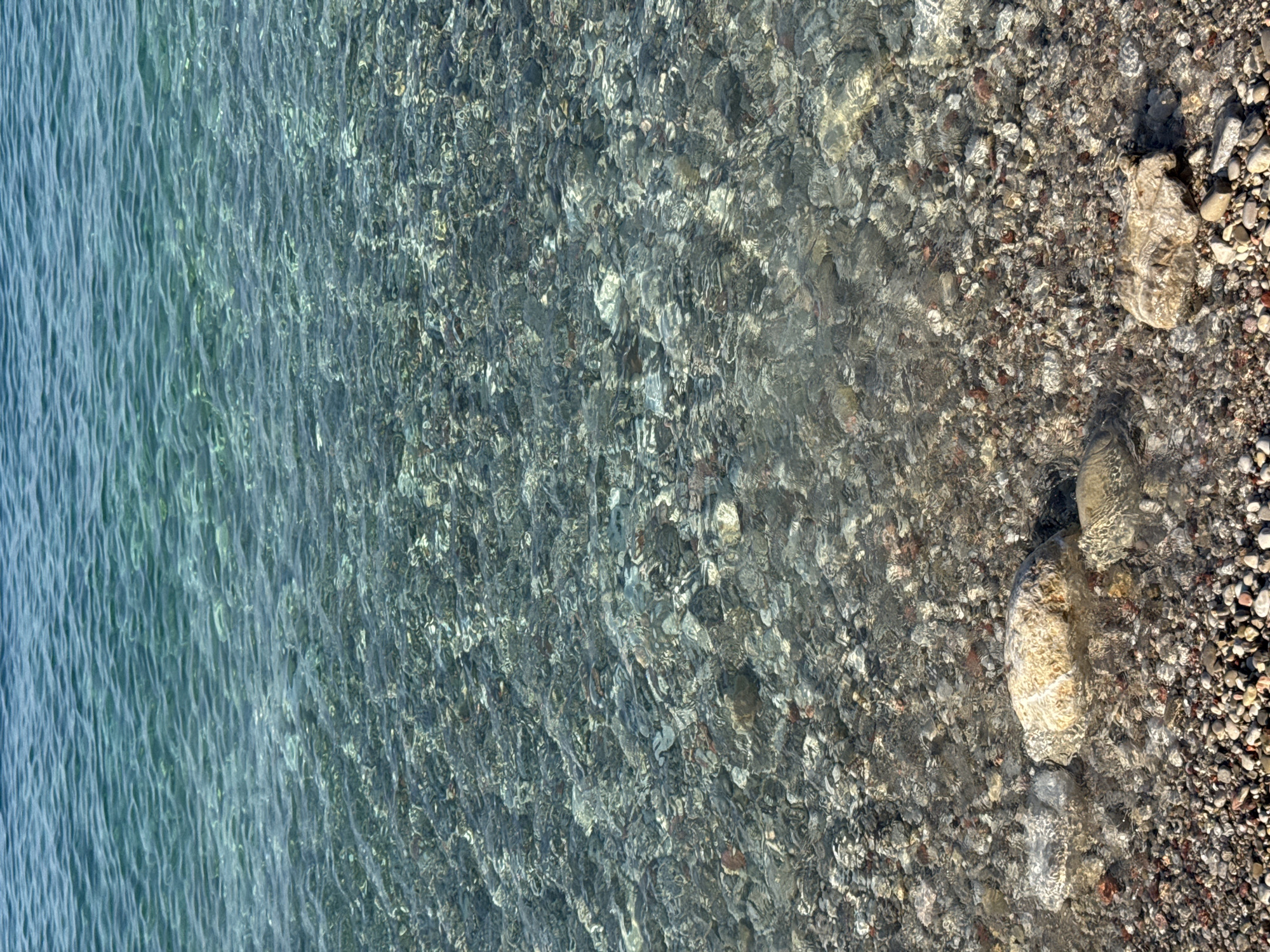The image depicts a clear, shallow body of water with a pebble-strewn seabed. The water exhibits a gradient of blue and green hues, with visible ripples on its surface. The seabed is composed of small, multicolored stones, with larger, lighter-colored rocks partially submerged in the water. The scene is an outdoor environment, likely a coastline or beach. There are no people or visible actions or interactions occurring in the image. The location is specified as Mirište, Montenegro.