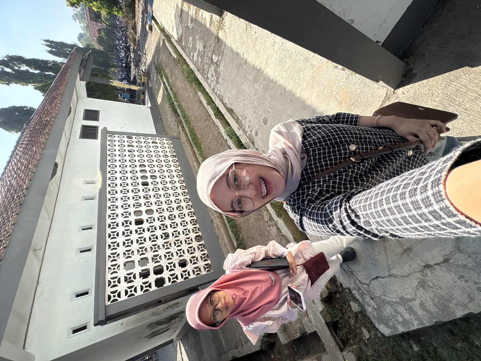 Two young women are taking a selfie outdoors. The woman in the foreground, closer to the camera, is wearing a pink hijab and glasses, and is smiling. She is holding a black clutch and a smartphone. Behind her, the second woman, wearing a light brown hijab and glasses, is also smiling. She has braces and is wearing a black and white checkered jacket. Both women are dressed in modest attire.
They appear to be on a paved pathway next to a building with a white facade and decorative white screens. The building has several small rectangular windows. Trees and a parked motorcycle area are visible in the background, suggesting an outdoor setting, possibly a school or public area in Gunungsari, Indonesia. The lighting indicates it is daytime, with sunlight casting shadows, suggesting a bright, possibly warm day. The overall mood is cheerful and casual.