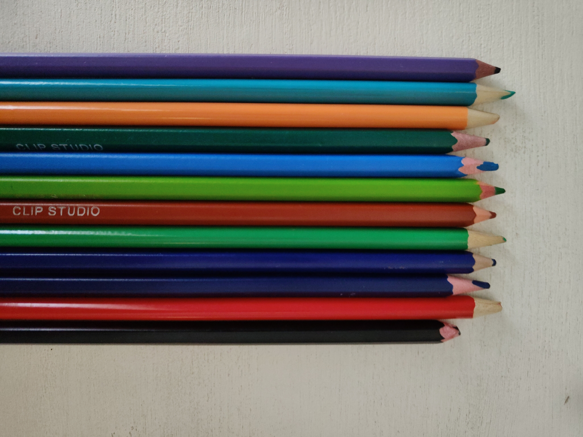 A top-down close-up photograph depicts a stack of ten colored pencils lying horizontally on a textured white surface. The pencils, arranged parallel to each other, exhibit a spectrum of colors from top to bottom: purple, teal, orange, dark green, blue, light green, brown, green, dark blue, red, and black. The points of the pencils are aligned on the right side of the frame. "CLIP STUDIO" is printed in white on two of the pencils. The scene's location is unspecified.