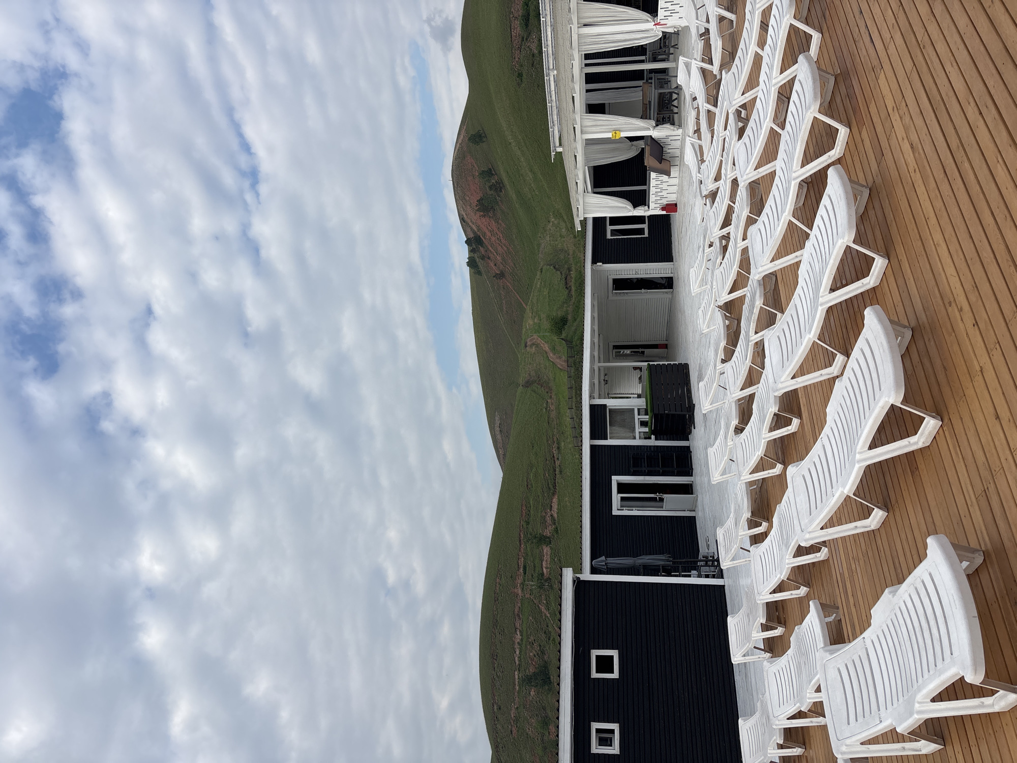 The image depicts an outdoor seating area at a resort or leisure facility. In the foreground, numerous white plastic lounge chairs are arranged on a wooden deck. Behind the lounge chairs, a low-rise building with black siding and white trim is visible. Several windows and doorways are present on this building. Further back, a white structure with white curtains is partially visible, suggesting a covered seating or dining area. The background features rolling green hills with some patches of reddish-brown soil and sparse vegetation. The sky is overcast with scattered clouds. The scene appears to be located in Drokino, Russia. No people are present in the image.