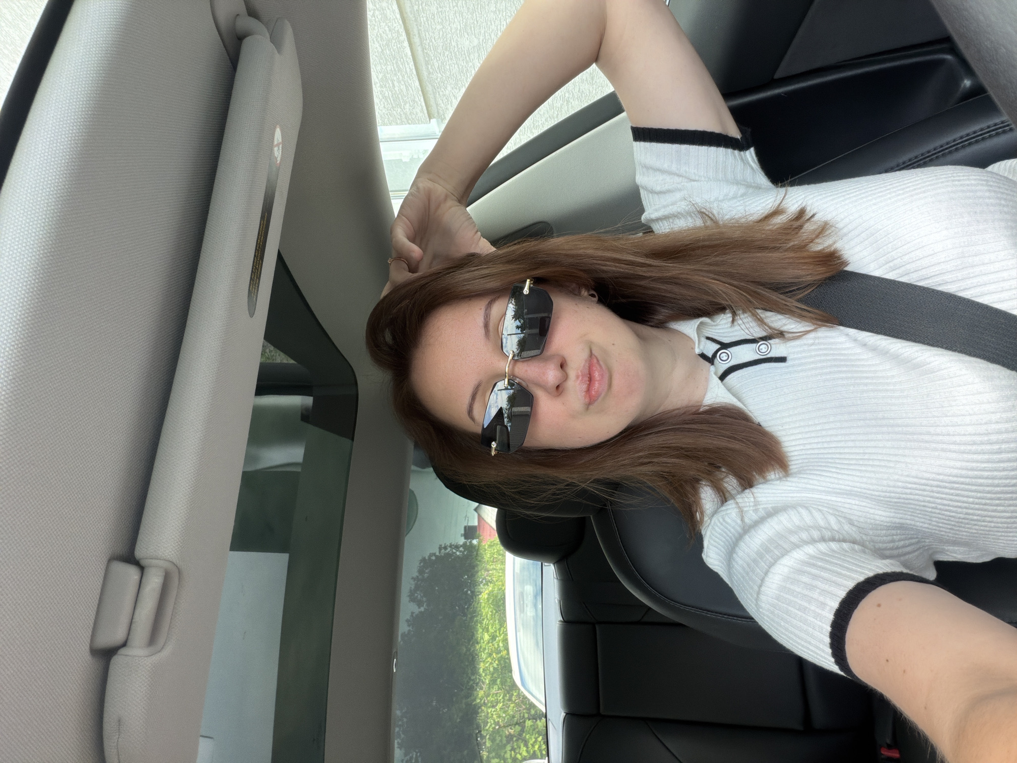 A young woman with brown hair and sunglasses is sitting in the driver's seat of a car, with her right arm raised and her hand behind her head. She is wearing a white collared shirt with black trim. The car has a sunroof. Outside the car, there is greenery and houses. The scene appears to be in Bila Tserkva, Ukraine.