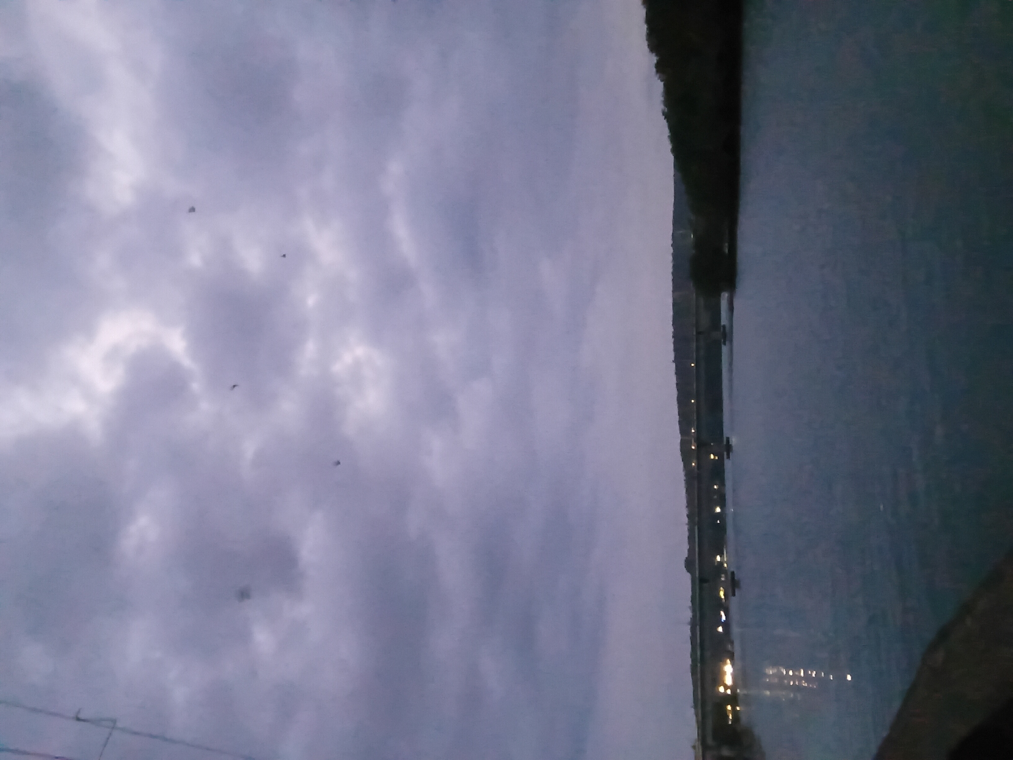 The image shows a body of water with city lights visible in the distance along the shoreline. On the left side of the image, a cloudy sky is visible. There are a few small dark objects, possibly birds, in the sky. The photograph is taken at dusk or dawn, based on the low light conditions. The location cannot be identified from the image.
