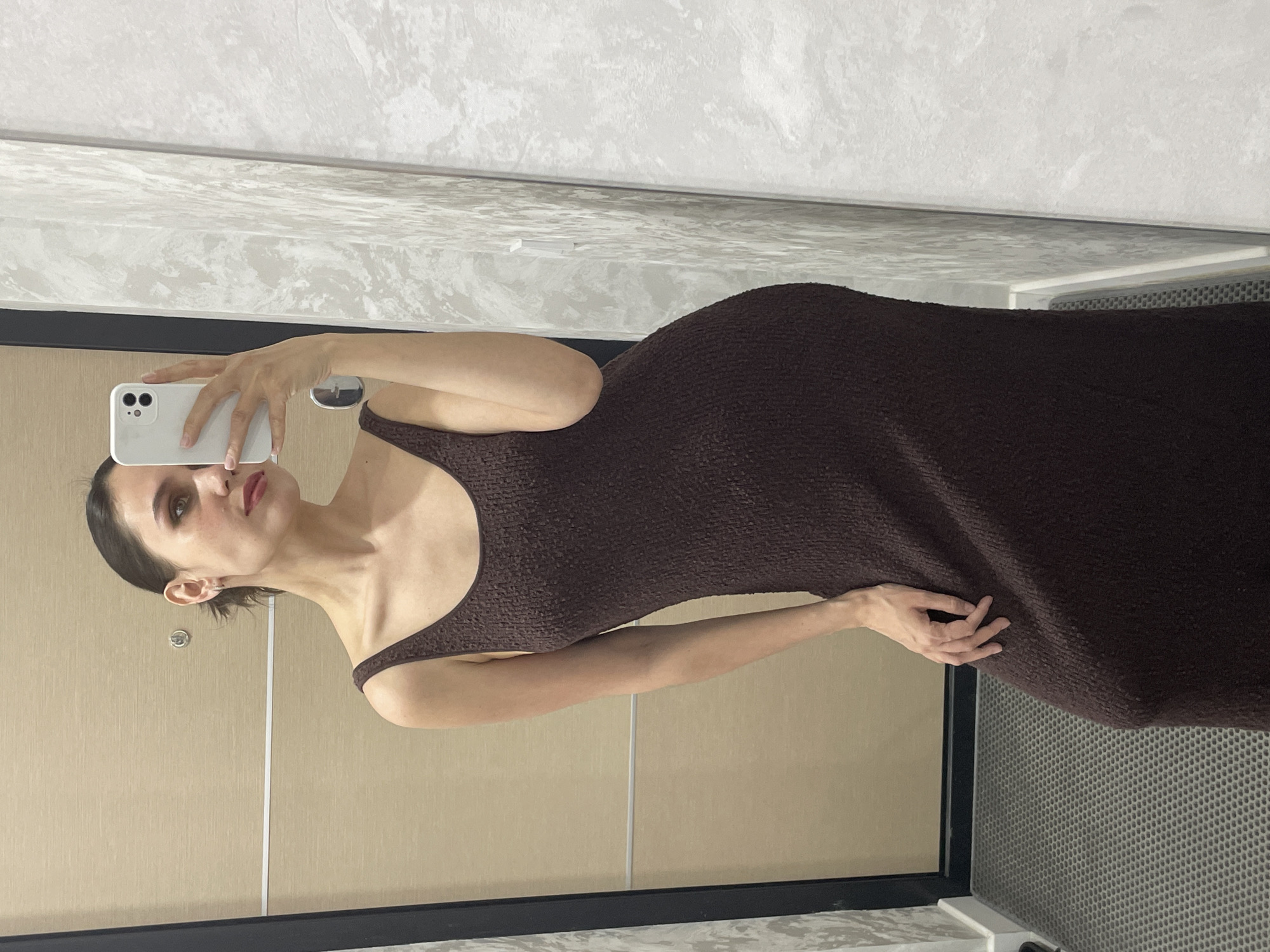 A woman in a brown, textured sleeveless dress is taking a selfie in a room. The phone is held up by her left hand. The room has a light beige or tan paneled wall and a white and gray patterned surface on the lower right, likely a floor or counter. The location is not identifiable as Izhevsk, Russia based on the provided image.